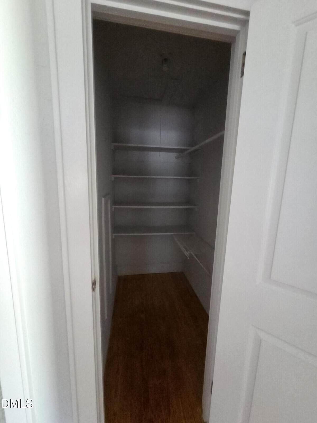 A small, empty walk-in closet with white shelves on the back and right walls, wooden floor, and an open white door.