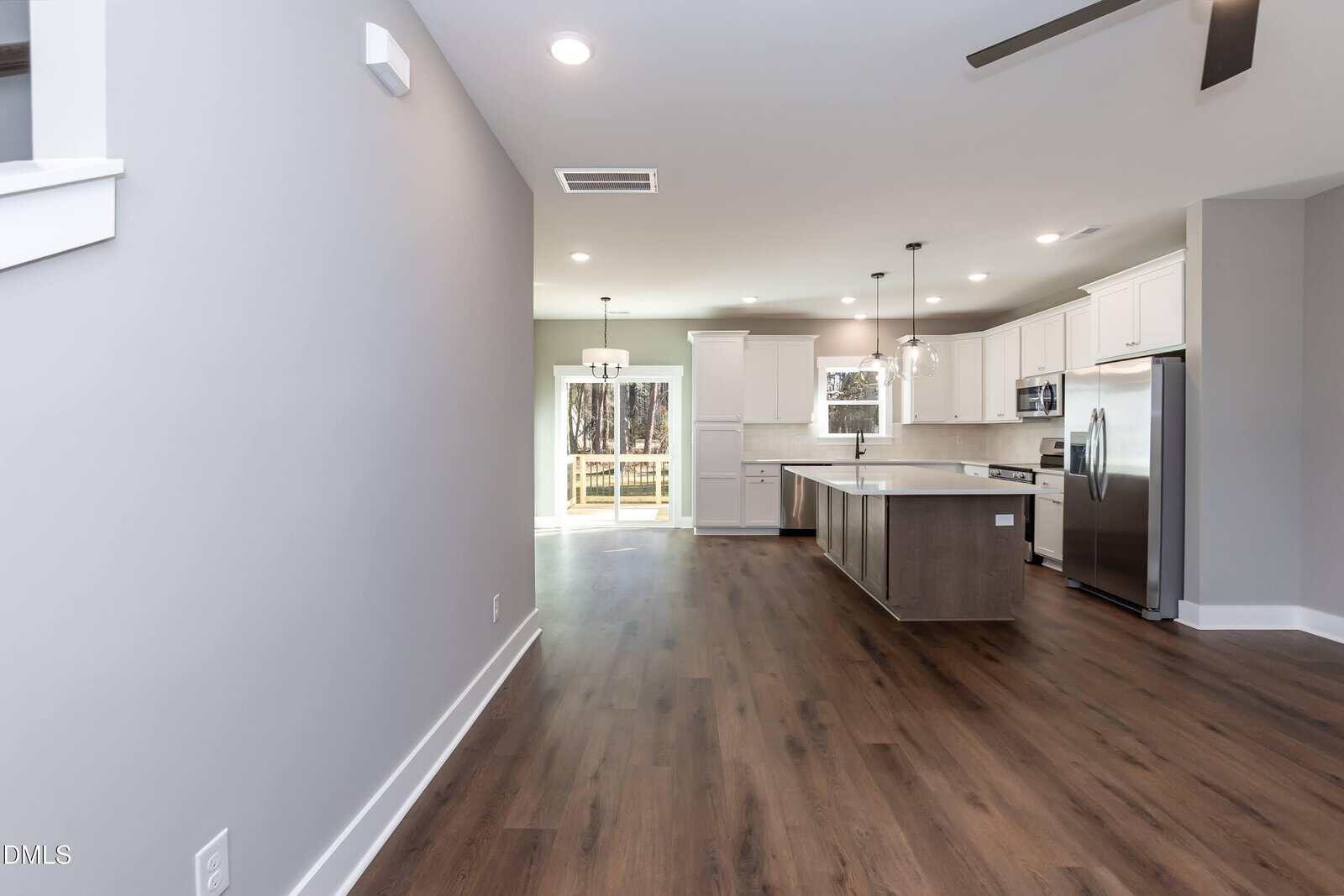 Open-plan kitchen with white cabinets, stainless steel appliances, a large island, pendant lights, and wood flooring. Glass doors at the back lead to a deck with trees visible outside. Neutral gray walls and ceiling fan.