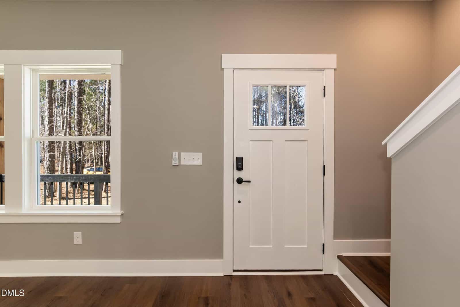 A modern entryway with a white front door, a window to the left showing trees outside, light taupe walls, white trim, wood flooring, and a staircase with a white banister on the right.