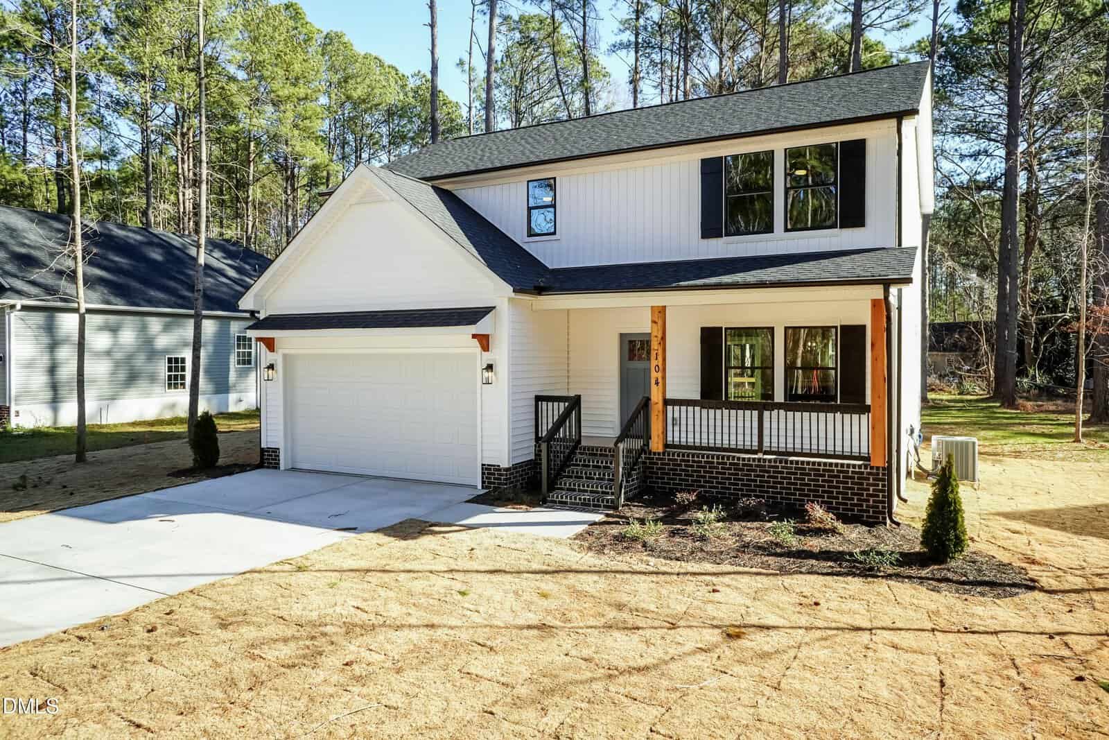 Modern two-story house with white siding, black roof, and a covered front porch. Attached two-car garage and driveway. Surrounded by trees and a patchy lawn, with neighboring houses visible.