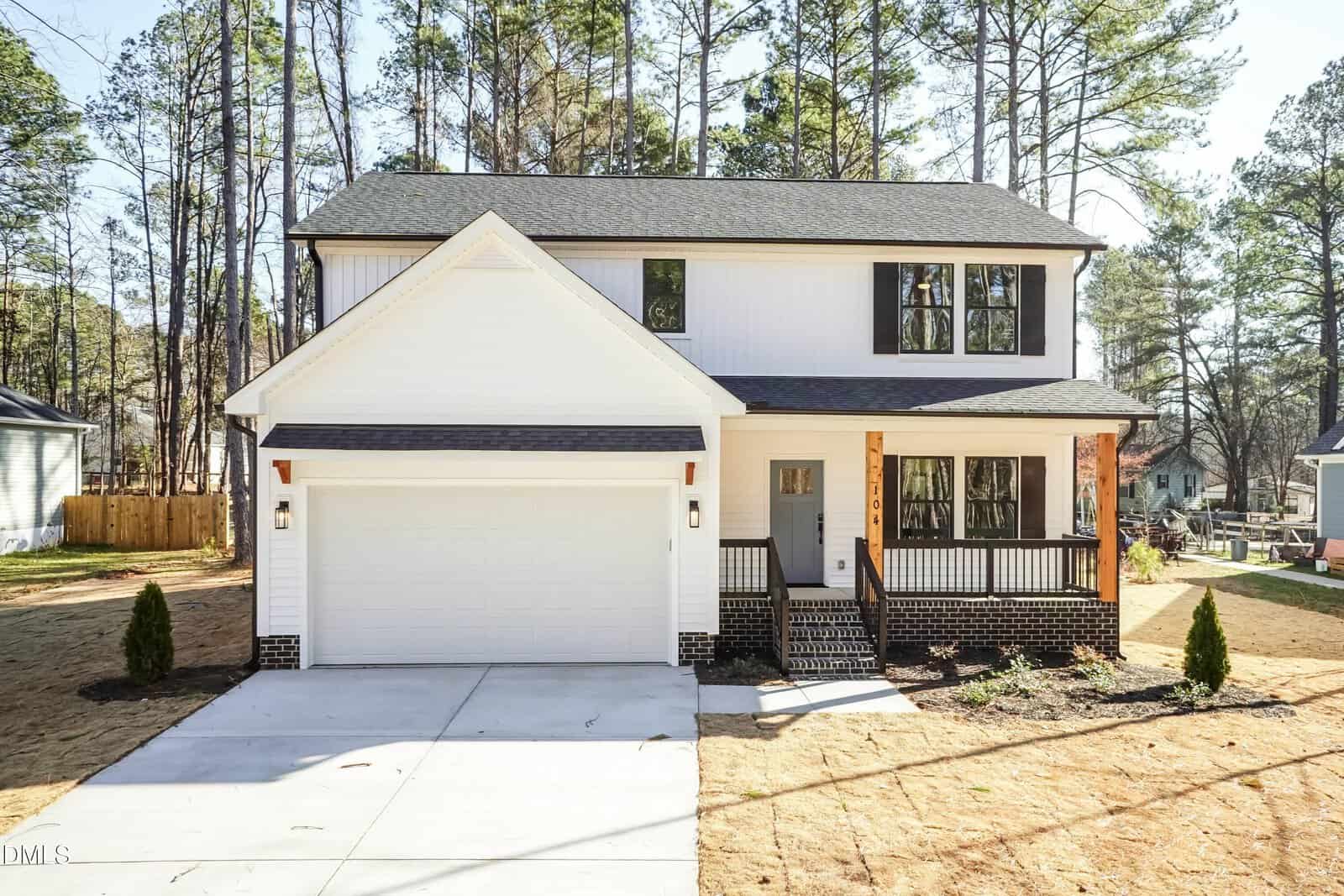 A modern two-story house at 104 Buffalo Drive, Louisburg, featuring white siding, black trim, a covered front porch, and an attached two-car garage. Young trees and fresh landscaping stand out against the scenic forested background.