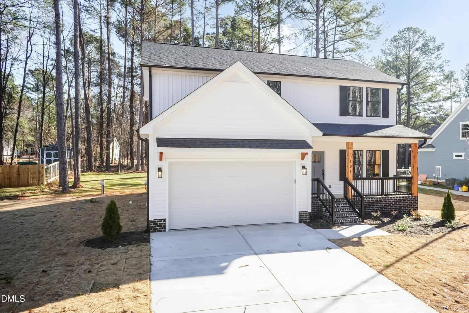 Two-story white house with black trim, a covered front porch, and a double garage. The home has a driveway, a small front yard with young trees, and is surrounded by tall pine trees.
