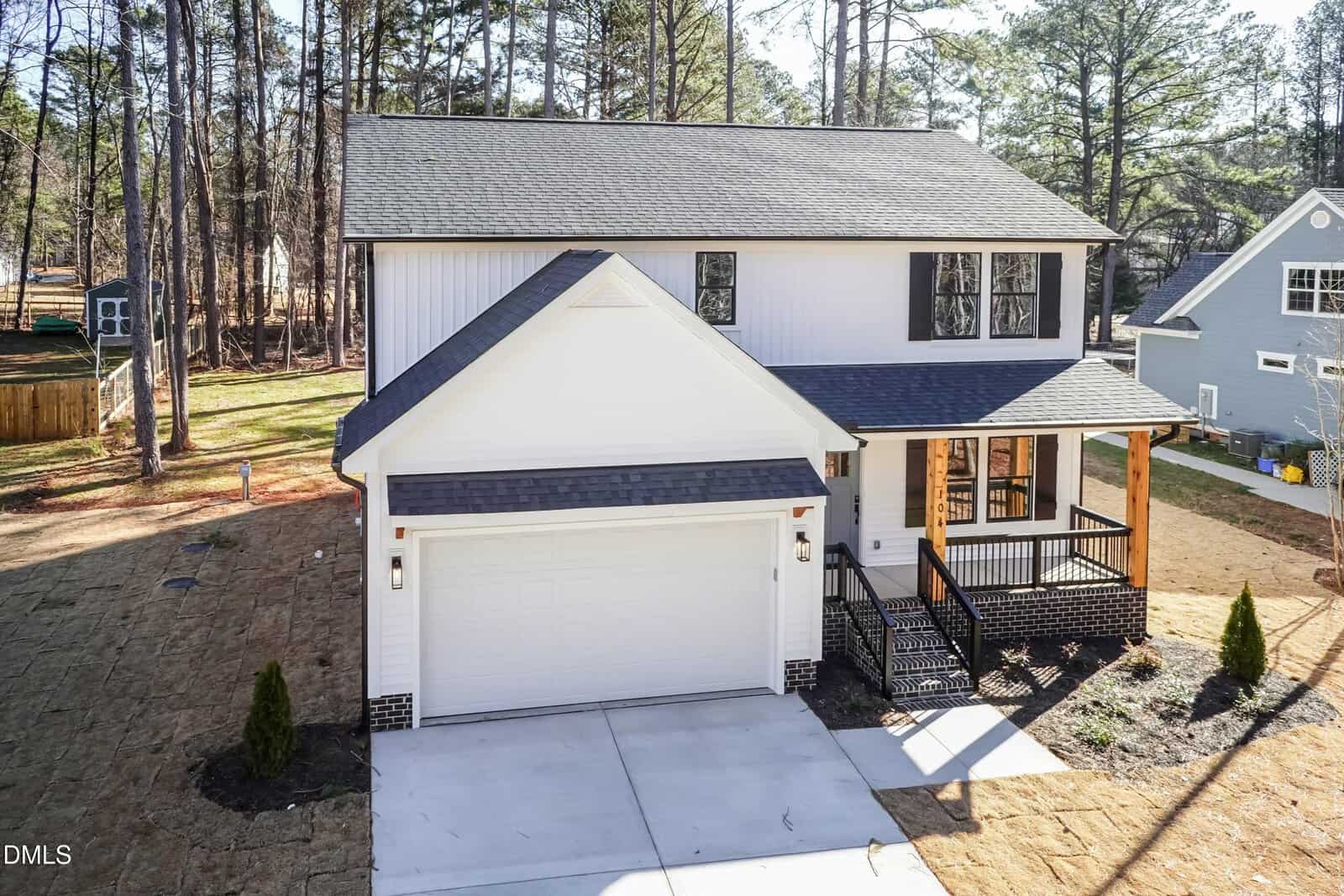 Two-story white house with black shutters, a covered front porch supported by wooden columns, and an attached two-car garage. The yard is partially landscaped, surrounded by tall trees and neighboring homes.