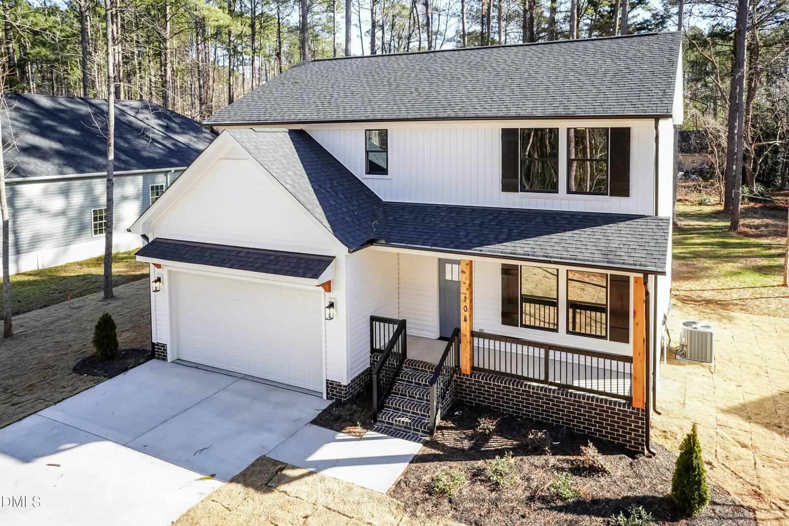 A two-story modern house with white siding, black trim, and a covered front porch. There is a double garage, paved driveway, and a small front yard surrounded by trees.