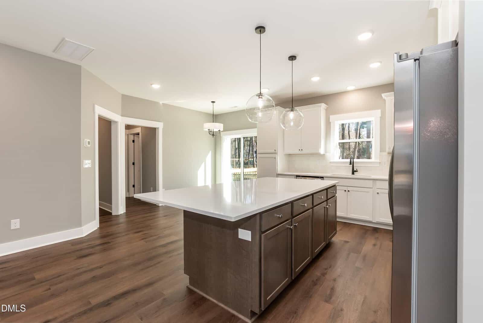 Modern kitchen with dark wood floors, large white island, stainless steel appliances, white cabinets, two round pendant lights, and windows letting in natural light. Walls are light gray. Dining area visible in background.