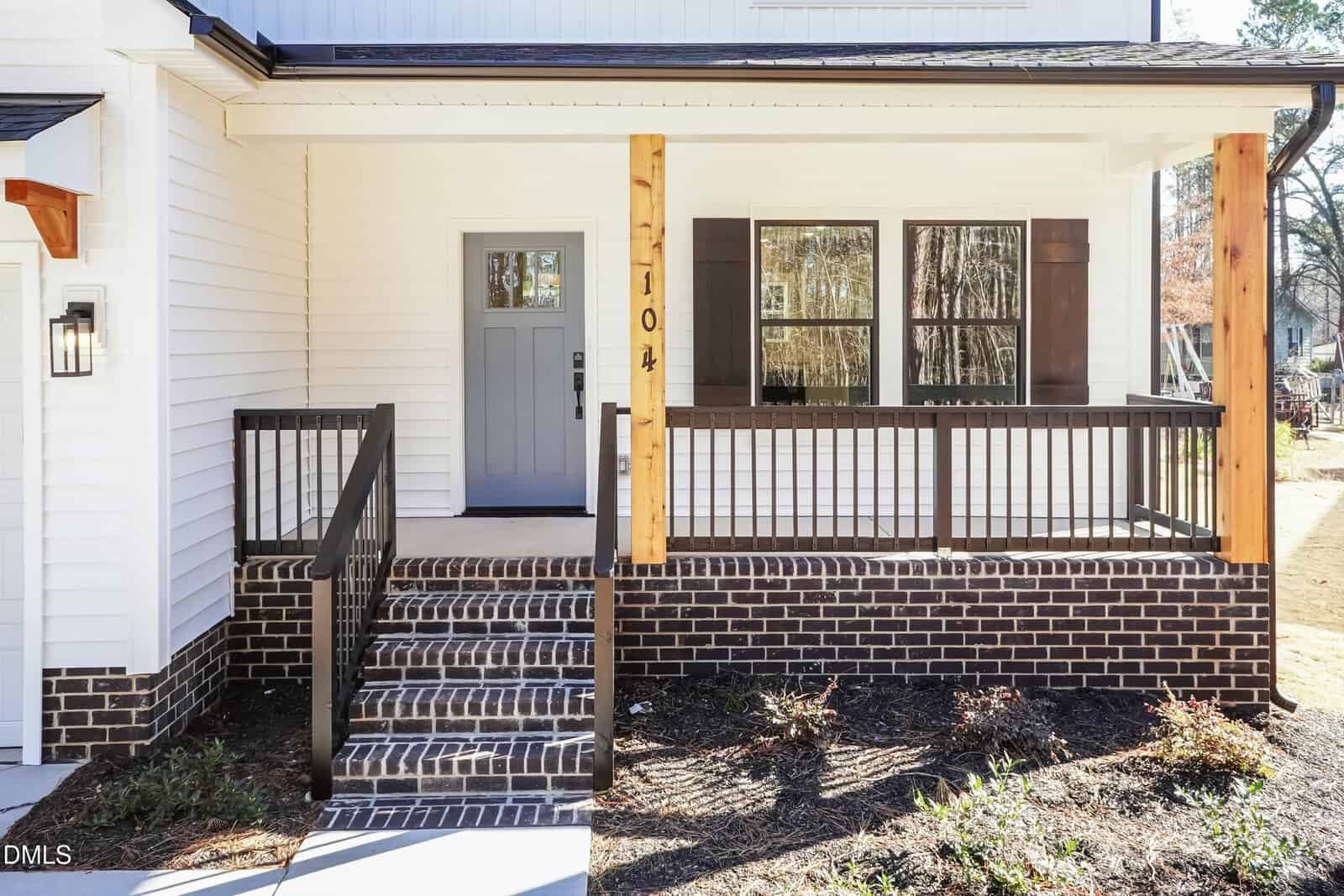 A modern white house with a gray front door, wooden posts, black railing, and brick steps leading up to a covered front porch. The yard is bare with some mulch and shadows from nearby trees.