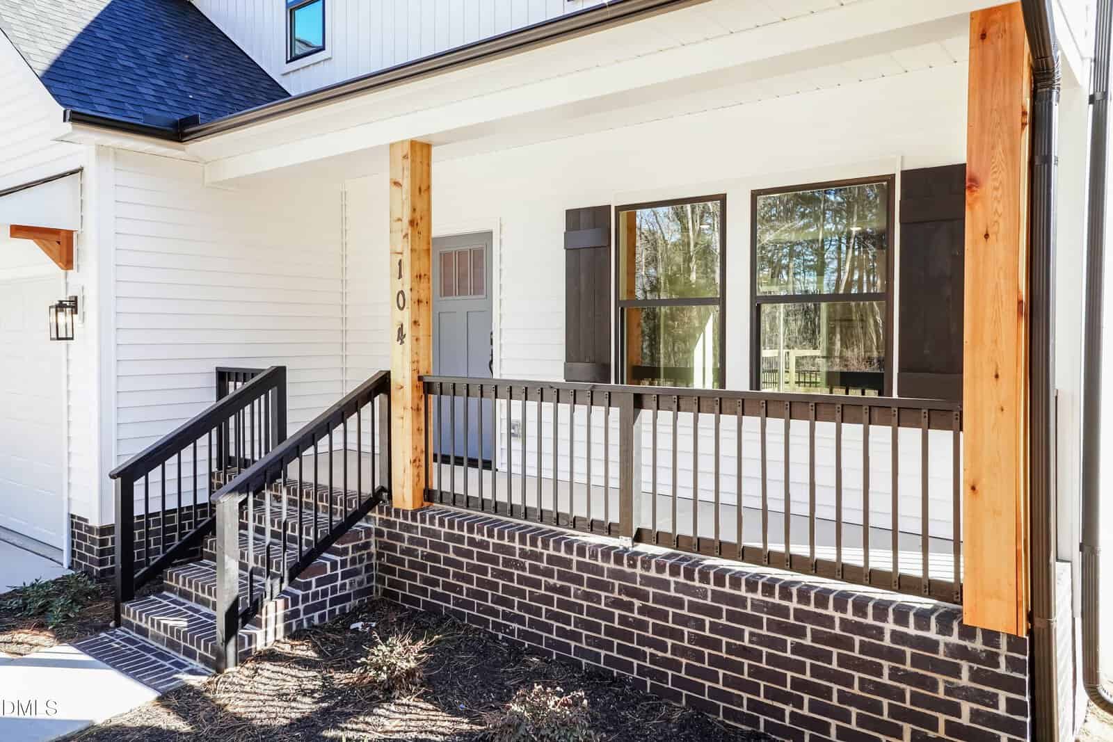 A modern house porch with white siding, black trim and railings, wooden posts, brick foundation, and a gray front door with glass panels next to large windows with black shutters.
