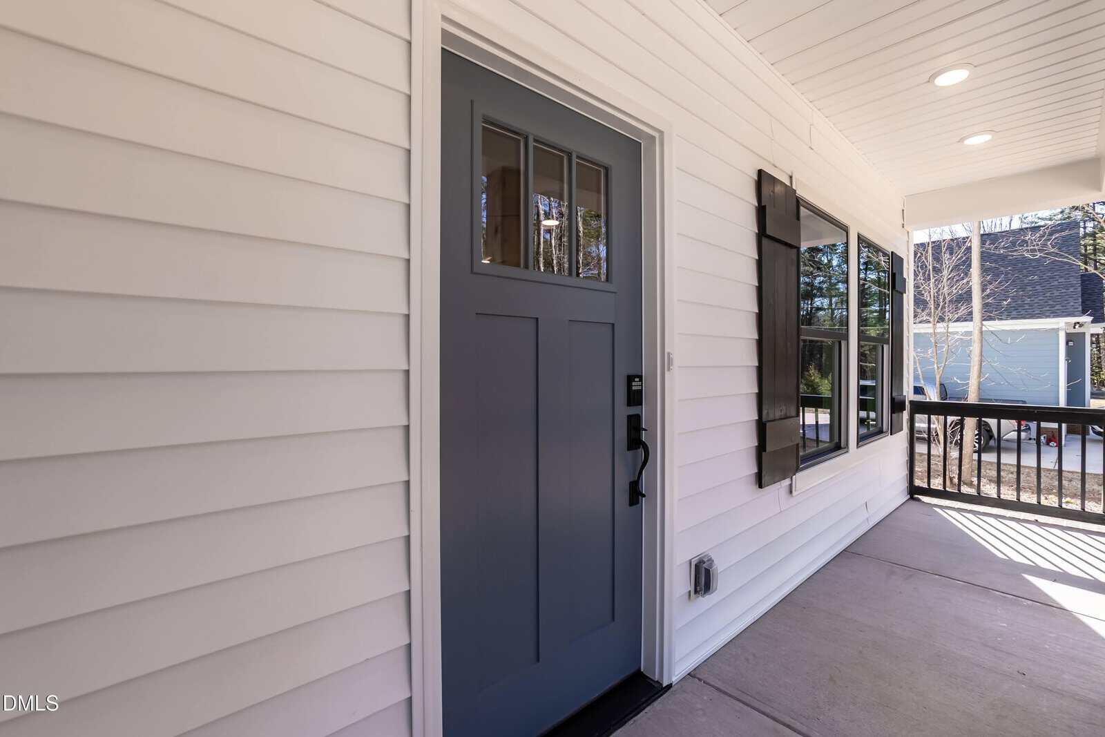 A close-up view of a blue front door with glass panels on a white house with siding. The porch has a black railing, and large windows with black trim are visible to the right of the door.