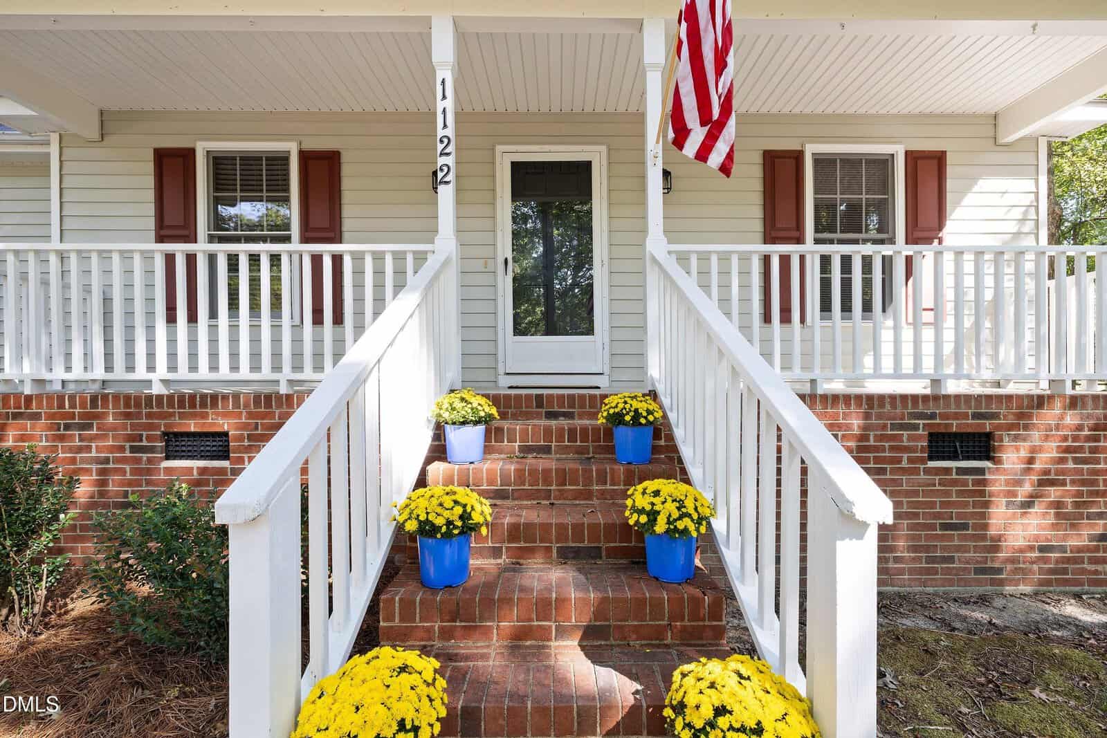 A brick porch with white railings leads to a front door. Six yellow potted flowers in blue pots line the steps. An American flag hangs on the right. The house exterior is white with red shutters.