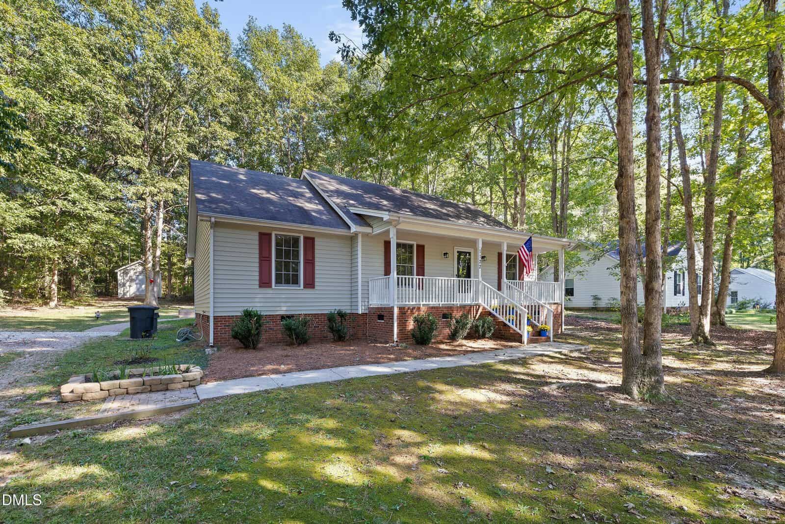 A single-story, light-colored house with a brick foundation, dark shutters, and a white front porch with an American flag. The home is surrounded by trees in a wooded lot with a paved walkway leading to the front steps.