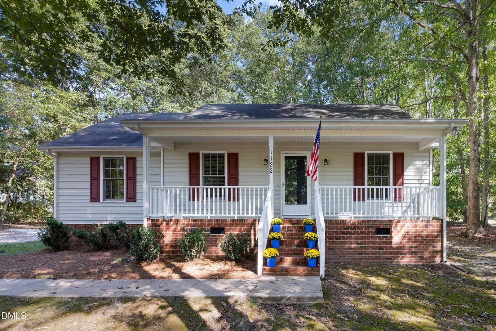 A single-story house with white siding, red shutters, a front porch, and brick steps decorated with yellow and purple potted flowers welcomes you at 1122 Country Club Lane in Zebulon. An American flag is displayed near the entrance, while trees surround the home.