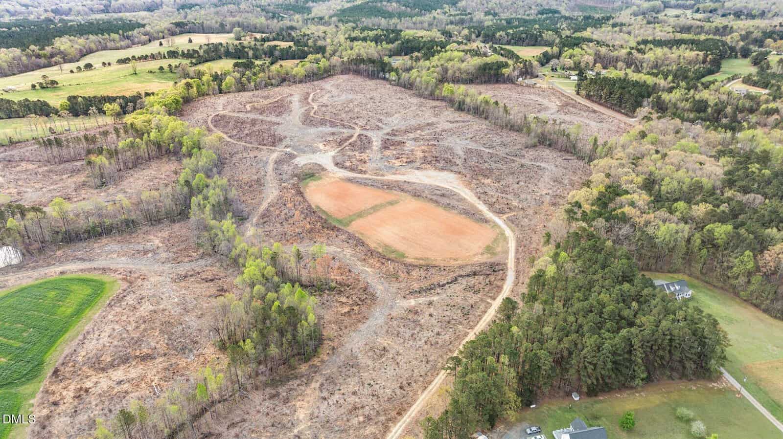 Aerial view of a large, partially cleared plot of land surrounded by trees, with winding dirt paths and a central cleared area bordered by forest and a few scattered houses.