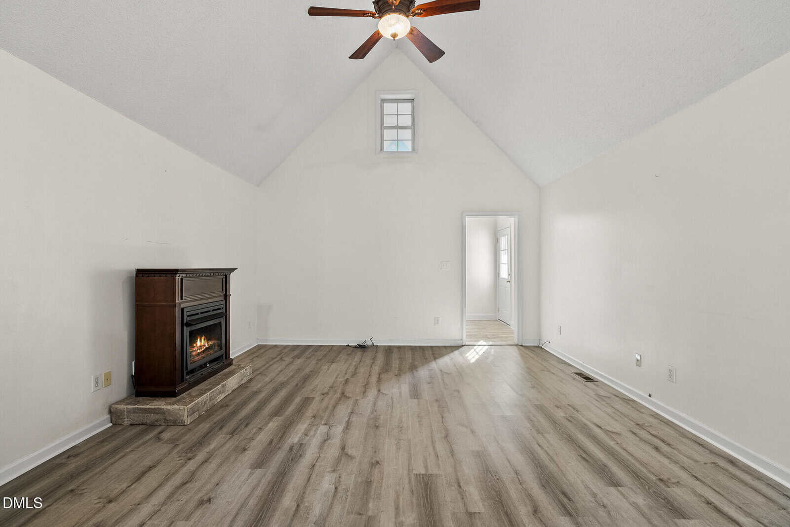 Unfurnished living room with vaulted ceiling, ceiling fan, light-colored walls, wood-style flooring, a small fireplace on a stone base, and a doorway leading to another room. Sunlight comes through a small window above the door.