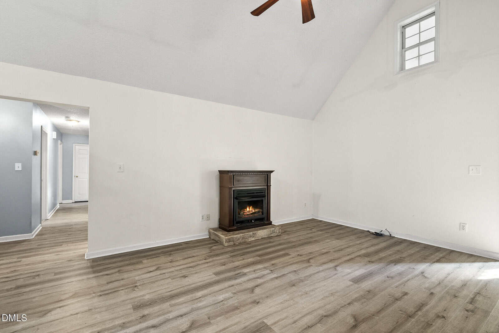 Unfurnished living room with light wood flooring, white walls, a high vaulted ceiling, ceiling fan, small fireplace on stone hearth, and a high window. A hallway and door are visible to the left.
