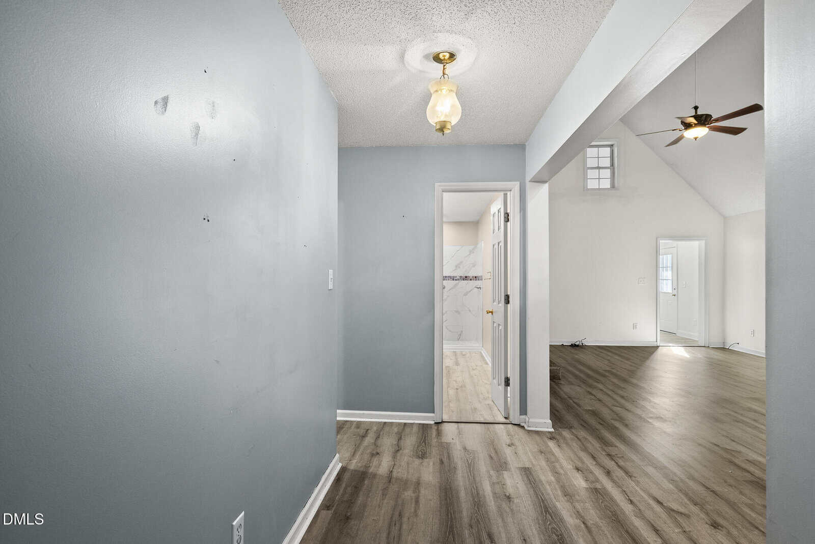 View of a hallway with gray walls and wood flooring, leading to a bathroom with white and marble tile. An open living area with vaulted ceilings, a ceiling fan, and large windows is visible to the right.