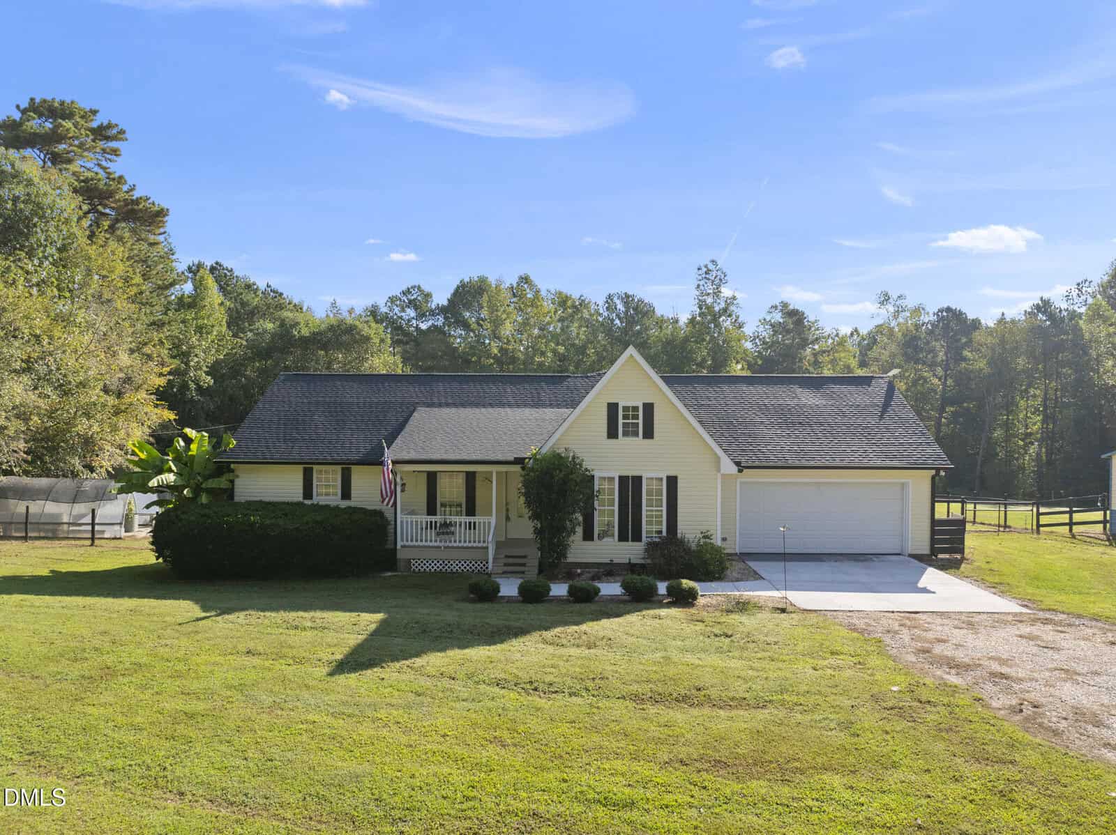 A yellow single-story house with dark roof, attached two-car garage, small front porch with white railing, American flag, and landscaped bushes, set on a grassy lawn with trees and blue sky in the background.