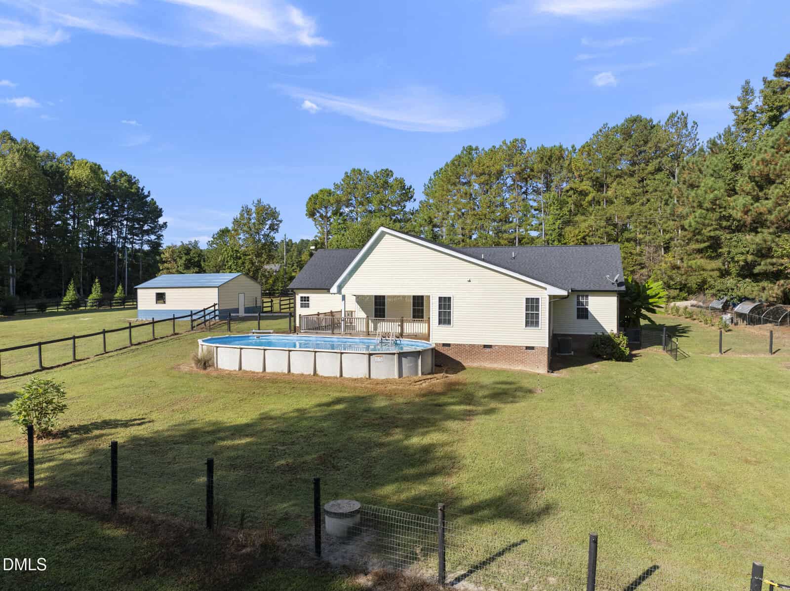 A single-story house with a dark roof sits in a large fenced yard, featuring an above-ground swimming pool and surrounded by trees. A separate small building is visible in the background.