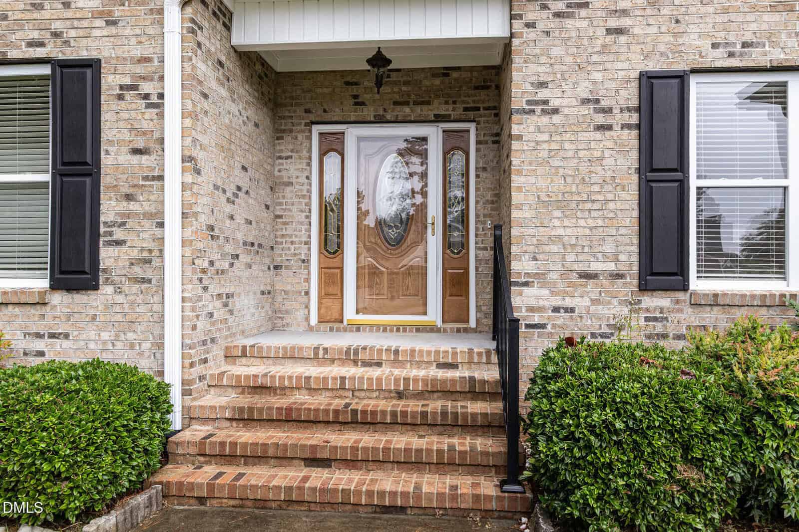 A brick house entryway with five brick steps, a wooden front door with glass panels, black shutters on both windows, and green bushes on either side of the steps.