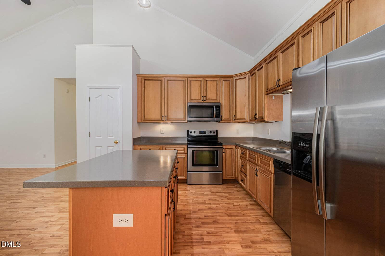 A modern kitchen with light wood cabinets, stainless steel appliances including a refrigerator, oven, and microwave, a gray countertop island, and hardwood flooring. White walls and high ceilings are visible in the background.