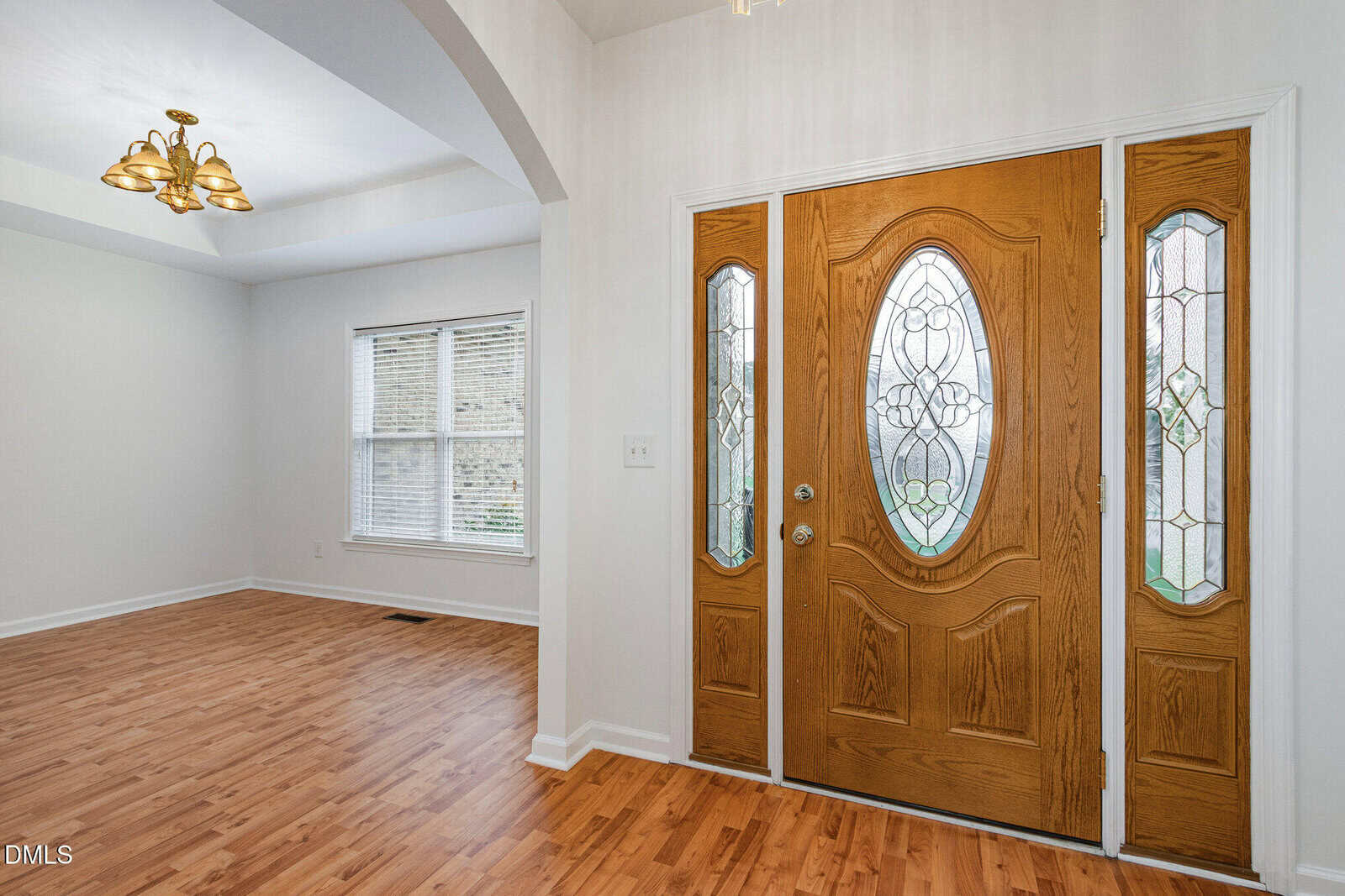 A wooden double front door with decorative glass panels opens into a home with light-colored walls and wood flooring. An adjacent room with a window and a brass ceiling light fixture is visible to the left.