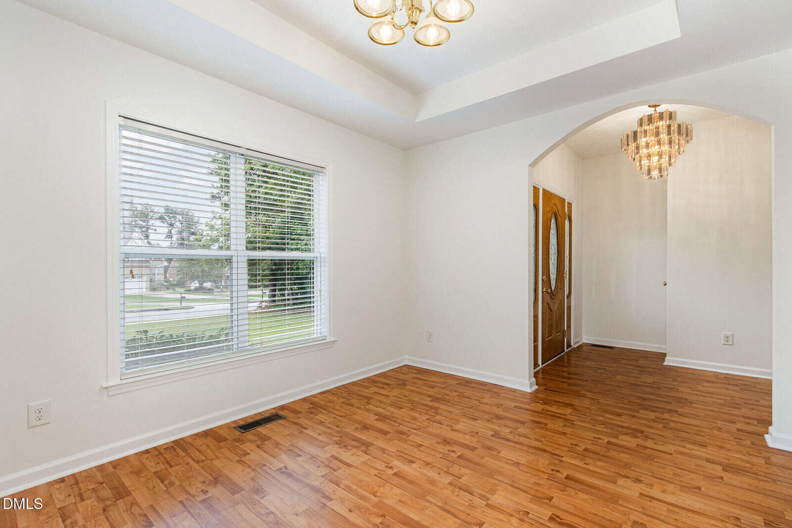 A bright room with wood floors, a large window with blinds, white walls, and a tray ceiling with a light fixture. An arched doorway leads to an entry area with a chandelier and a wooden front door.