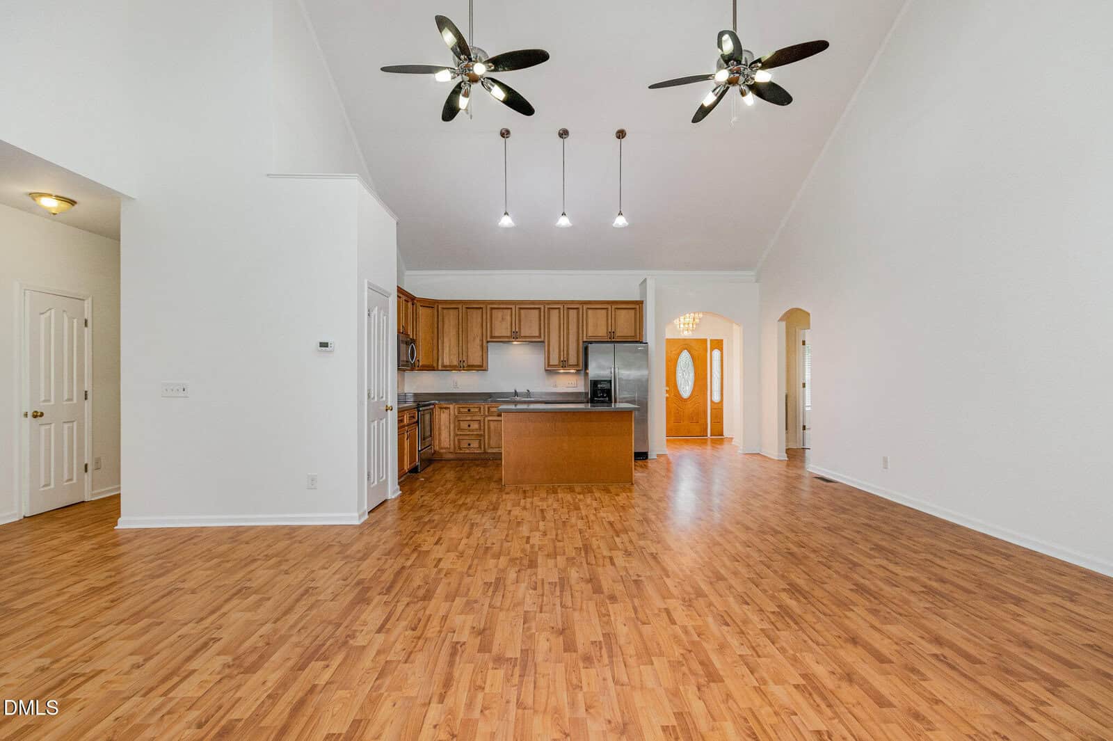 Spacious open-concept kitchen and living area with wood flooring, wood cabinets, an island, high ceilings, three black ceiling fans, and pendant lights. White walls and an arched entryway are visible.