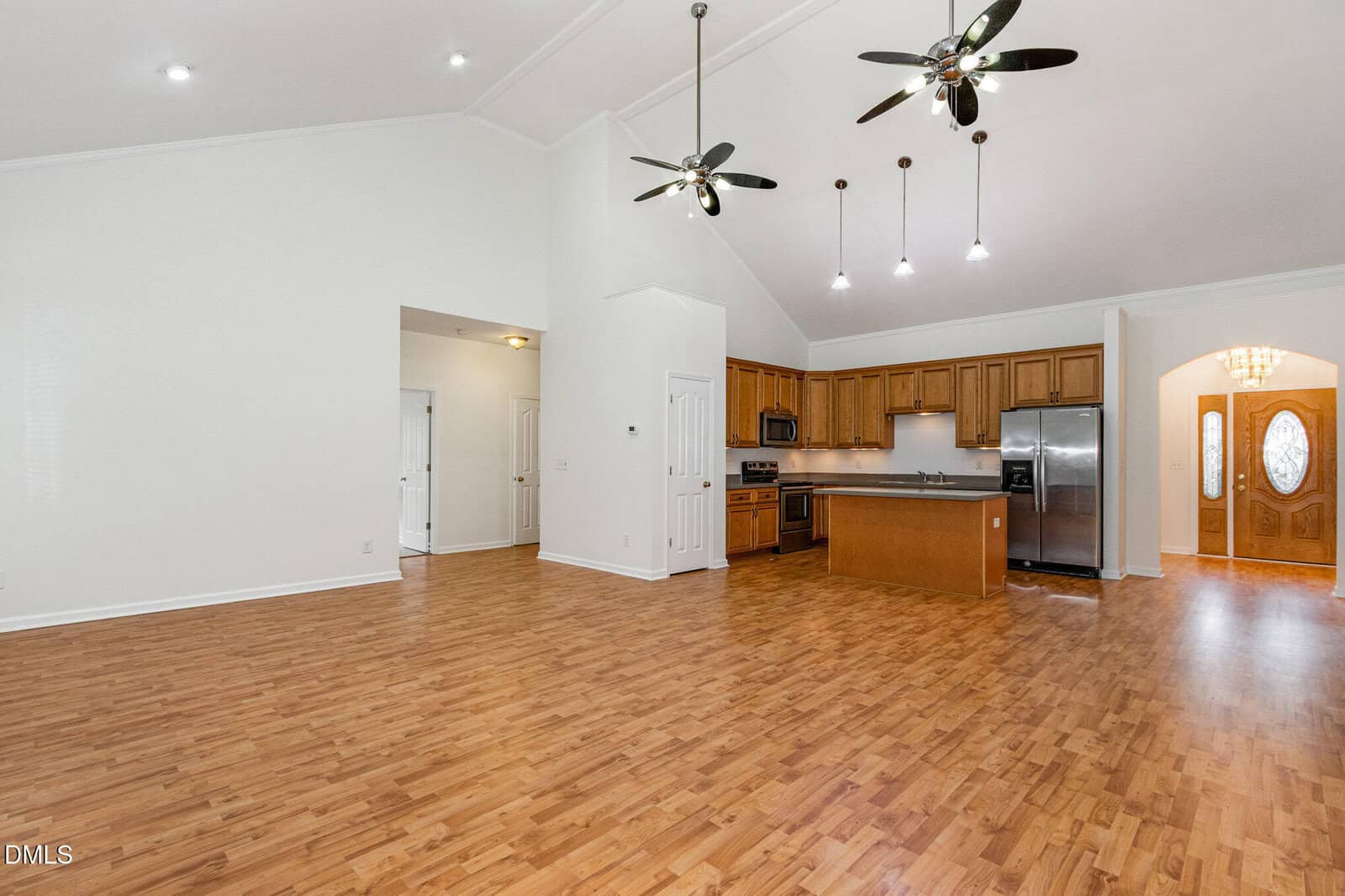 Spacious open-concept room with wood floors, high vaulted ceiling, two ceiling fans, a kitchen with wooden cabinets, stainless steel appliances, and an island; entryway with double doors visible in the background.