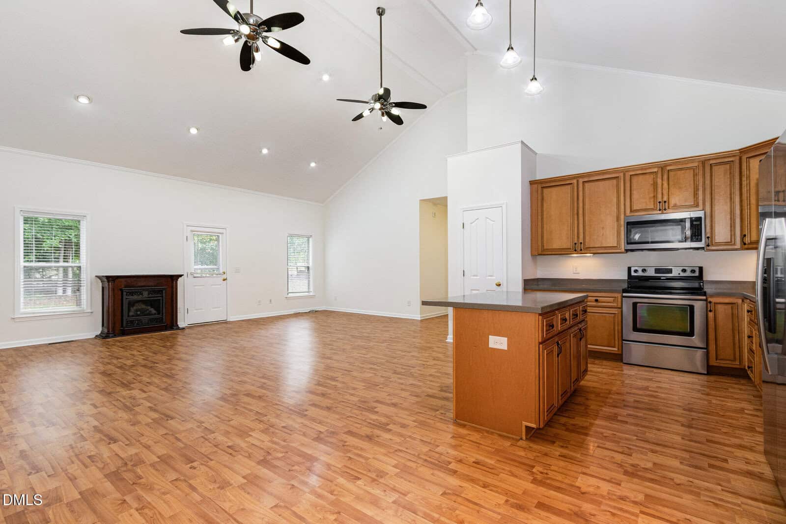 Spacious open-concept living area with wood flooring, high ceilings, ceiling fans, a fireplace, and large windows. The kitchen has wooden cabinets, stainless steel appliances, and a center island.