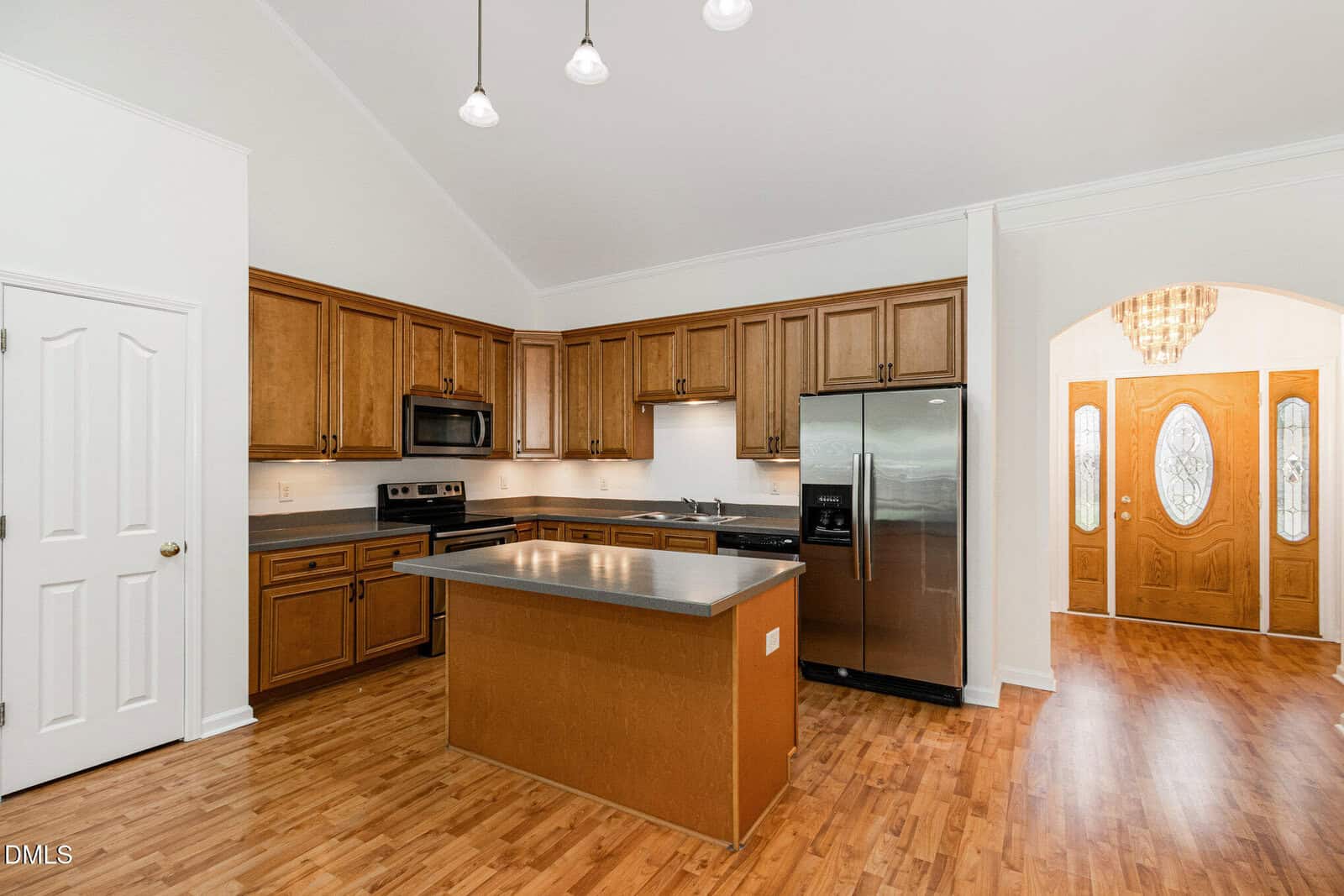 A kitchen with wood cabinets, stainless steel appliances, a center island, and hardwood floors. There is a white pantry door to the left and a view of the front entryway with double doors and a chandelier to the right.