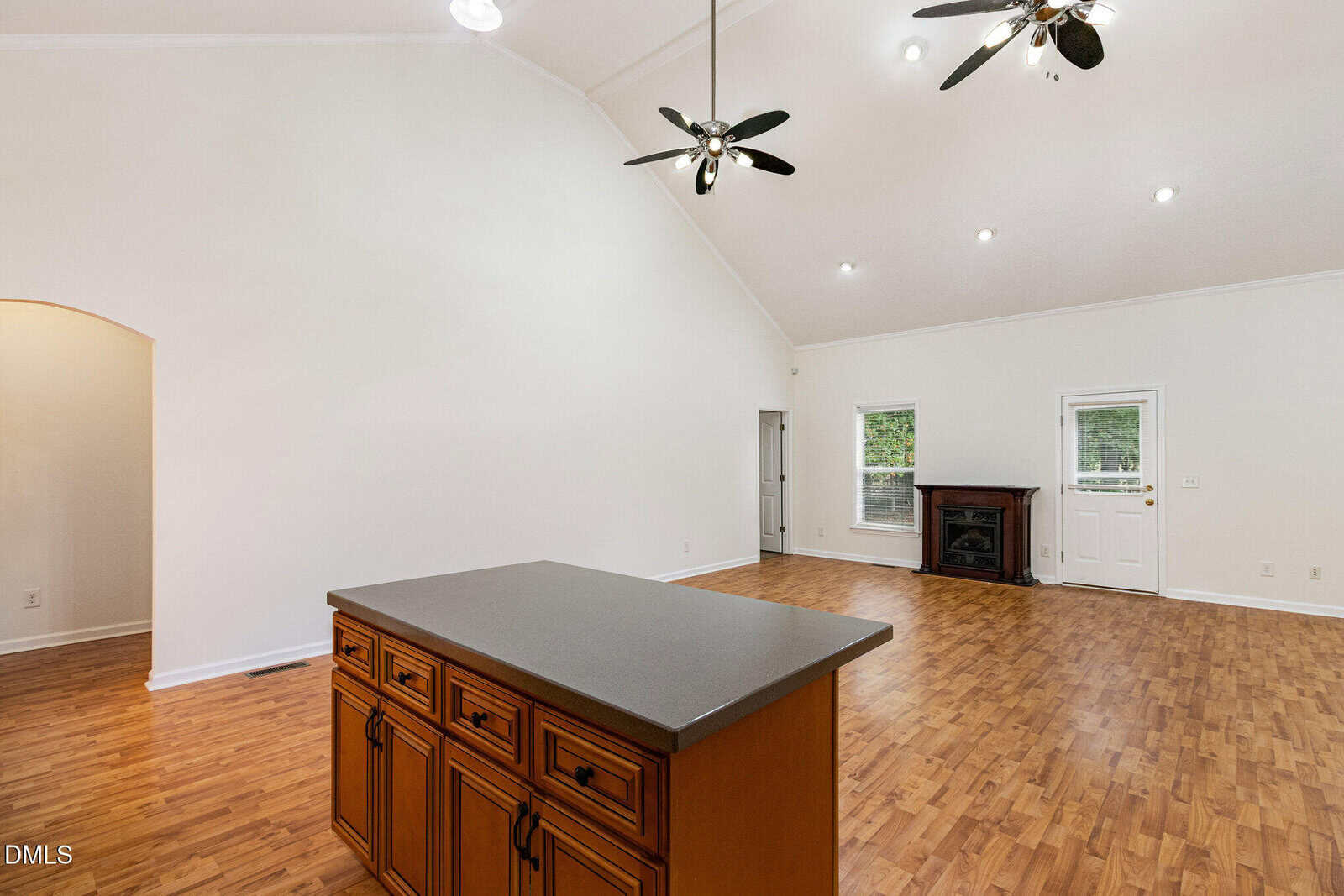 Spacious living area with high ceilings, ceiling fans, wood floors, an island with drawers, fireplace, and white walls. Large windows and a door let in natural light.