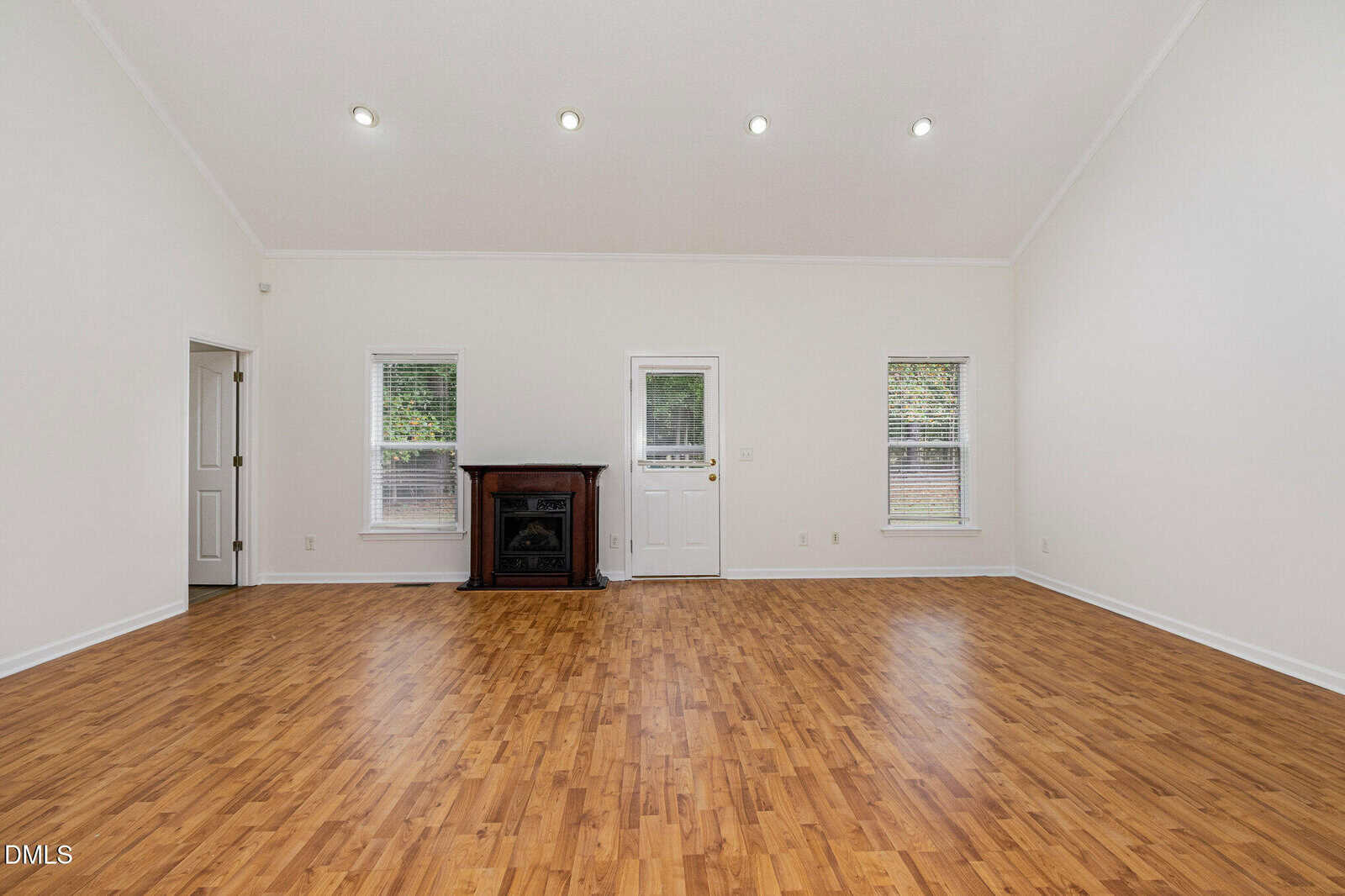 Spacious empty room with wood flooring, white walls, high ceiling, recessed lighting, two windows, a door, and a black fireplace centered between the windows.
