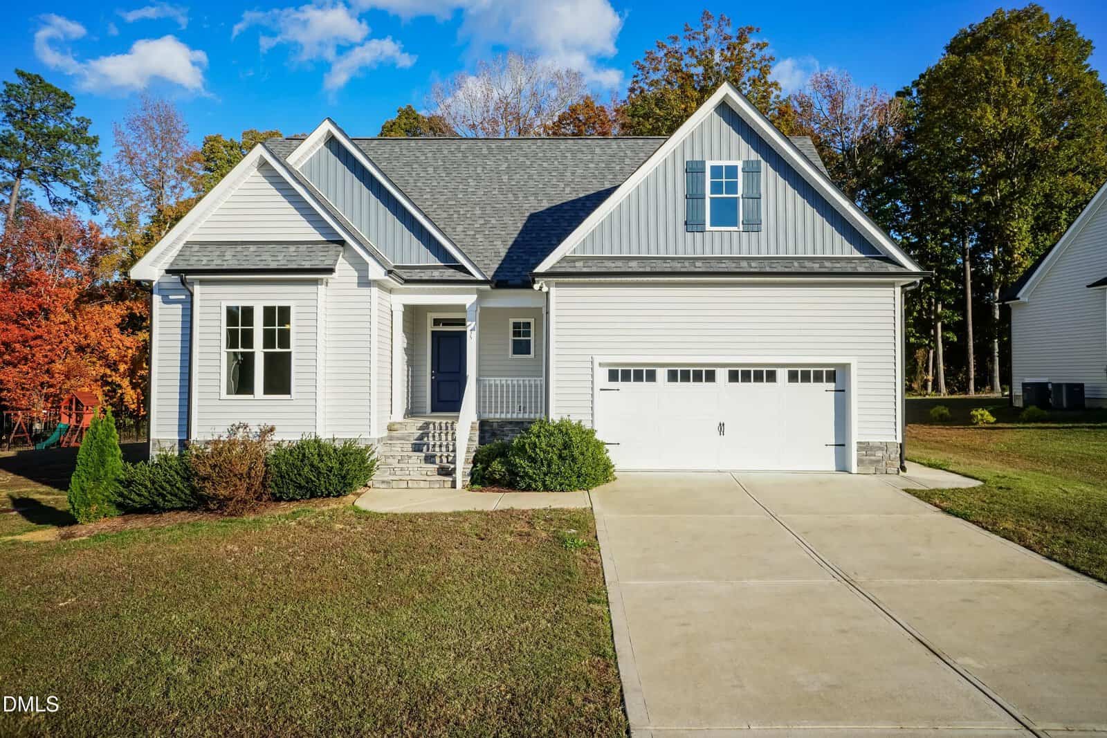 A modern gray and white single-family house with an attached double garage, front steps, small shrubs, and a wide driveway. Green grass and trees with autumn leaves surround the house. The sky is blue with some clouds.