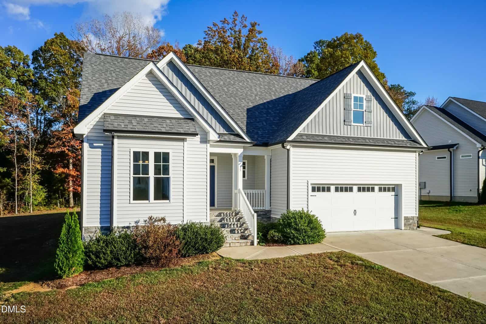 A modern single-family house with white siding, gray roof, double garage, front porch, and steps leading to the entrance. The lawn is partially grassed, with shrubs in front and trees in the background under a blue sky.