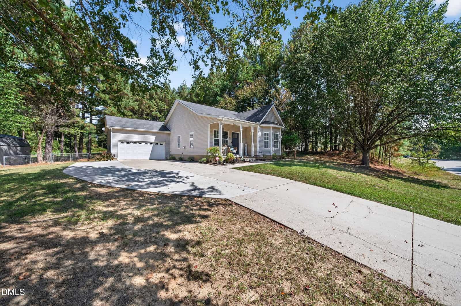 Single-story beige house at 25 Hillside Village Drive, Louisburg, featuring a welcoming front porch with white columns and an attached two-car garage. The property boasts a curved driveway framed by sparse grass and mature trees, all basking in the glow of a bright, sunny day.