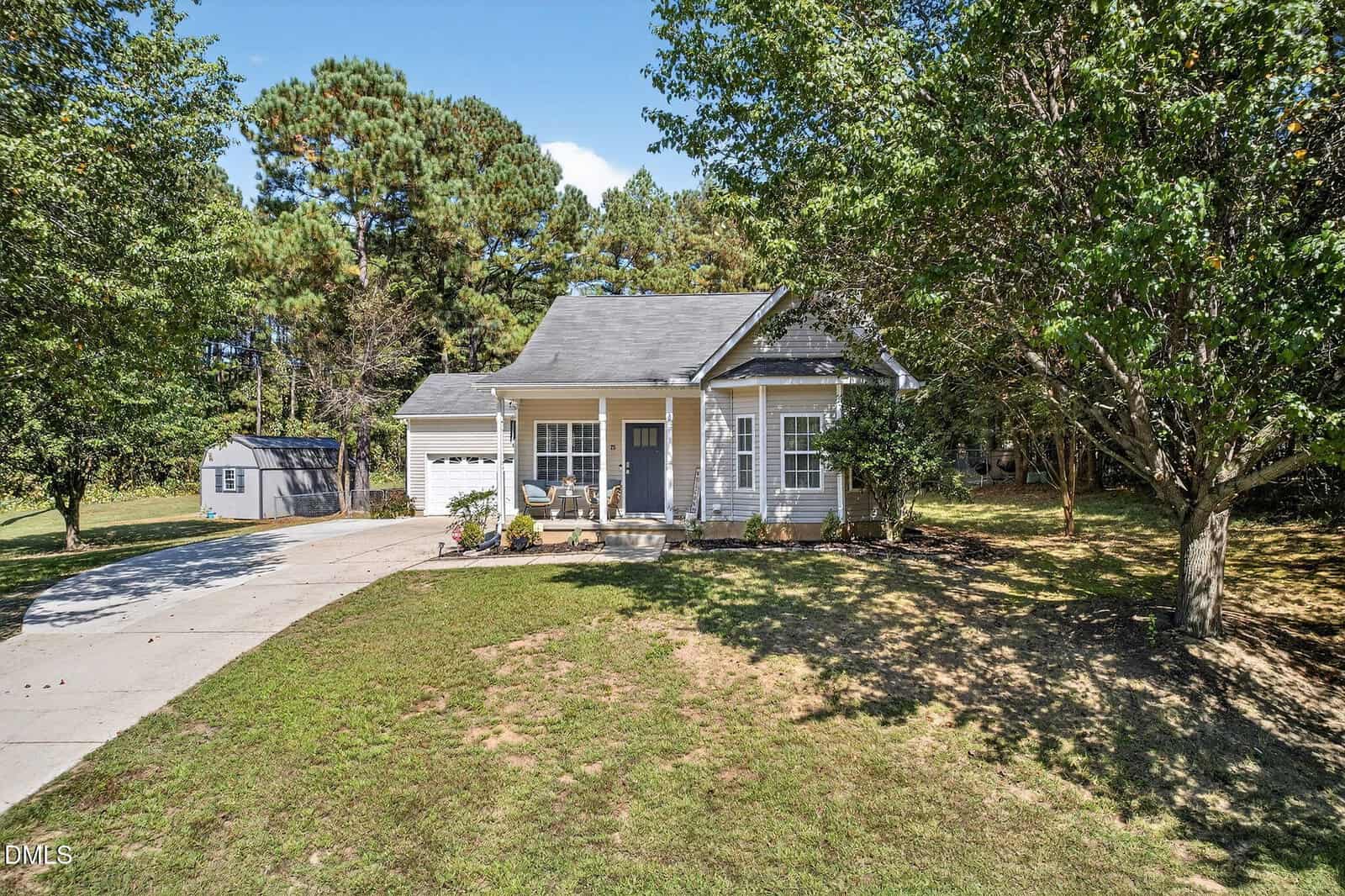 Single-story house with gray siding, front porch, attached garage, and a curved driveway. There is a lawn with some trees and a small shed in the background. The sky is clear and sunny.