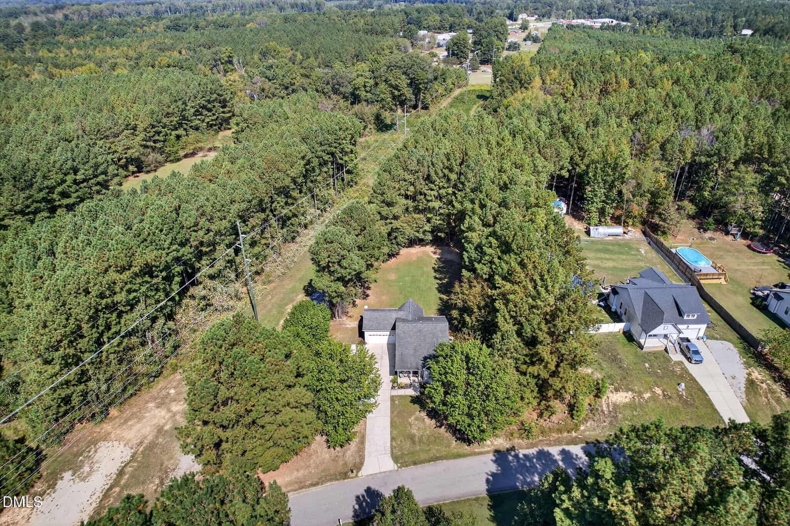 Aerial view of two houses surrounded by dense trees, with driveways leading to a road. One house has a pool in the backyard. The landscape is green and rural with forests in the background.