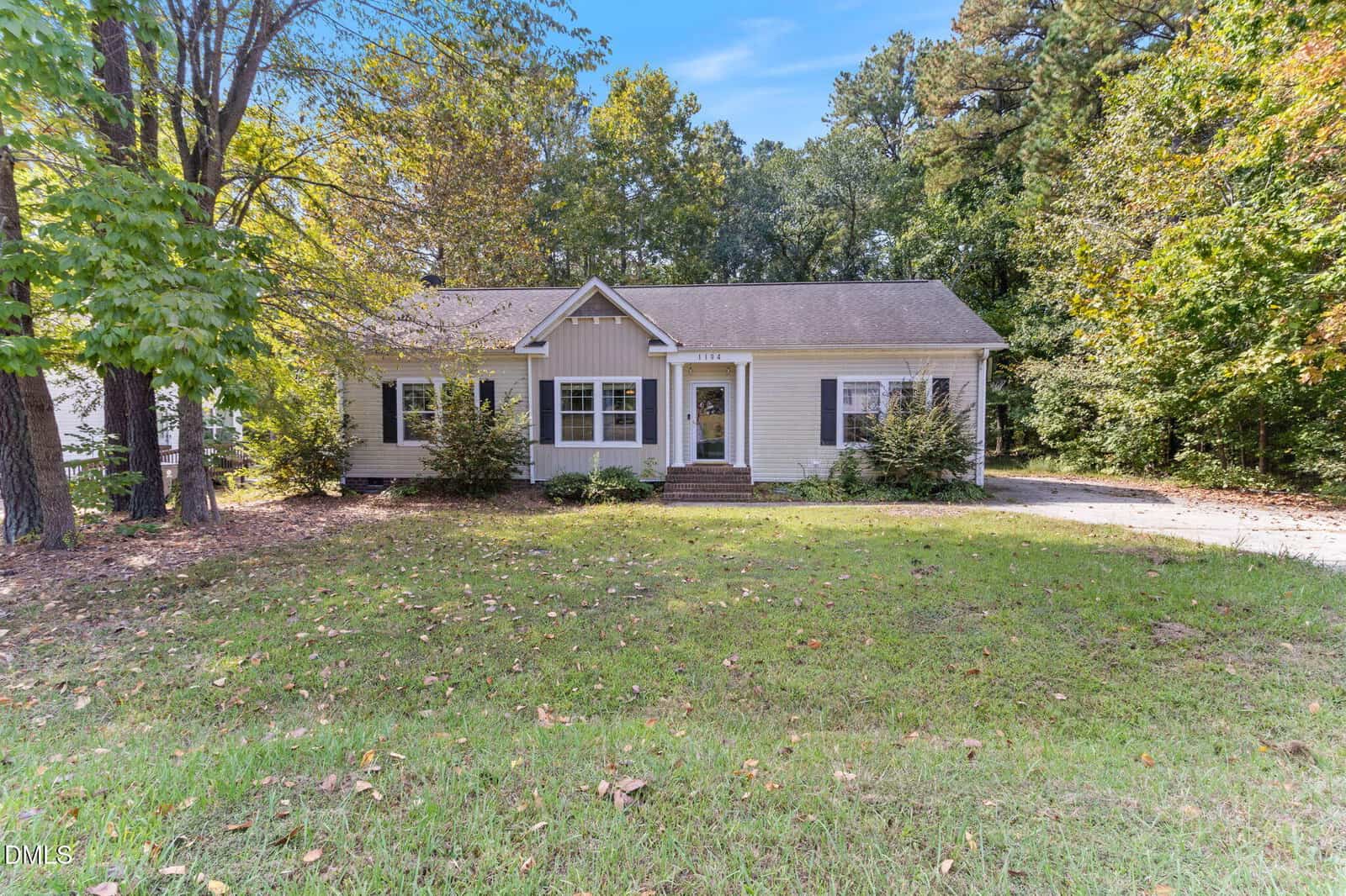 Nestled among trees in Creedmoor, 1194 E Middleton Drive is a single-story beige house with white trim, a gabled roof, and a cozy front porch. Shrubs flank the entrance, while fallen leaves scatter across the lawn and driveway on the right.
