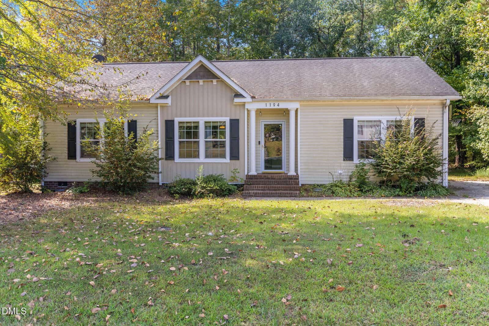 Single-story beige house with white trim and dark shutters, featuring a central front door with small stoop and steps, surrounded by green grass, bushes, and trees in the background.