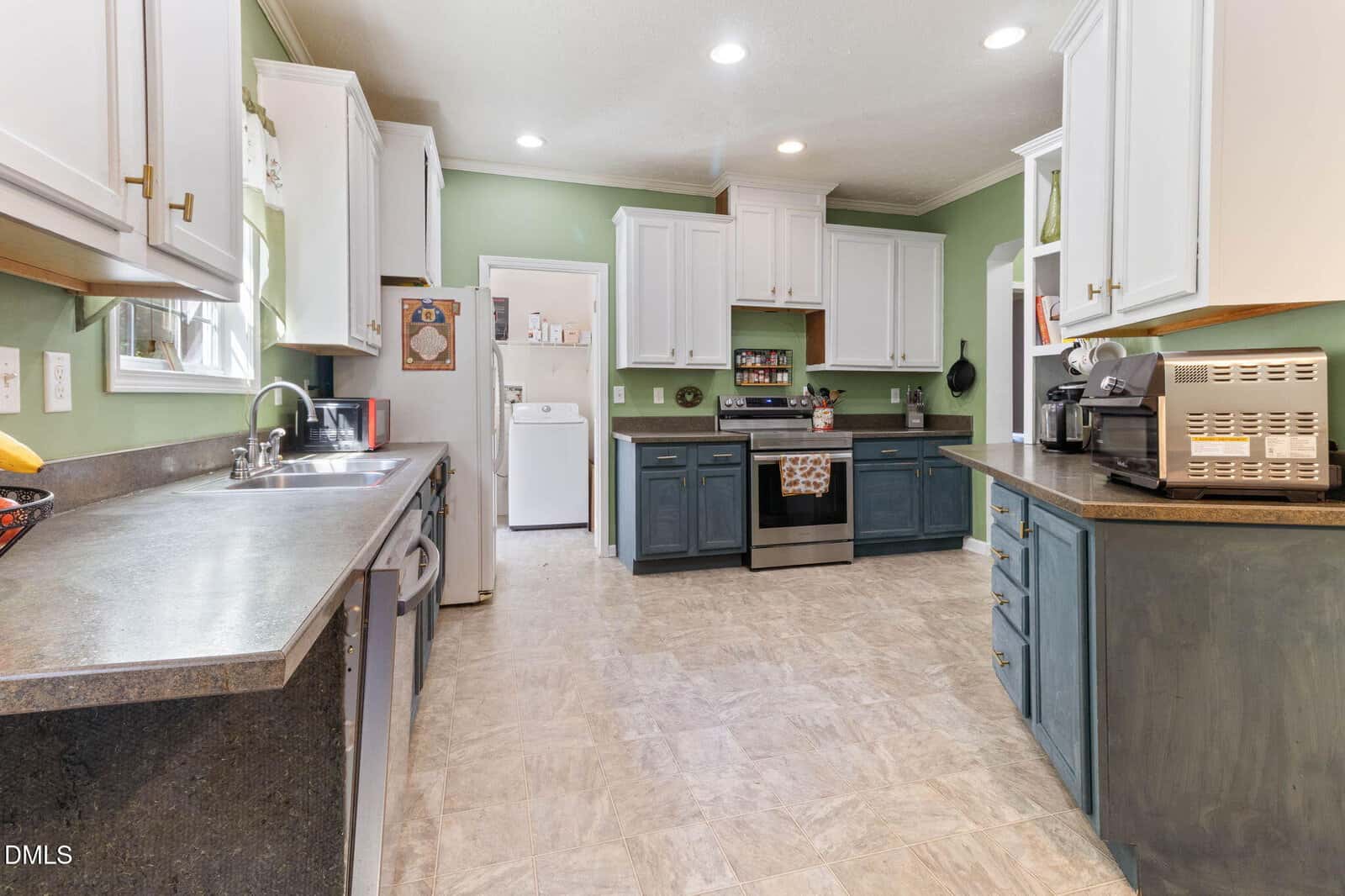 Spacious kitchen with green walls, white and blue cabinets, stainless steel appliances, and a view into a laundry room. Countertops hold a coffee maker, microwave, sink, and fruit. Recessed lighting brightens the room.