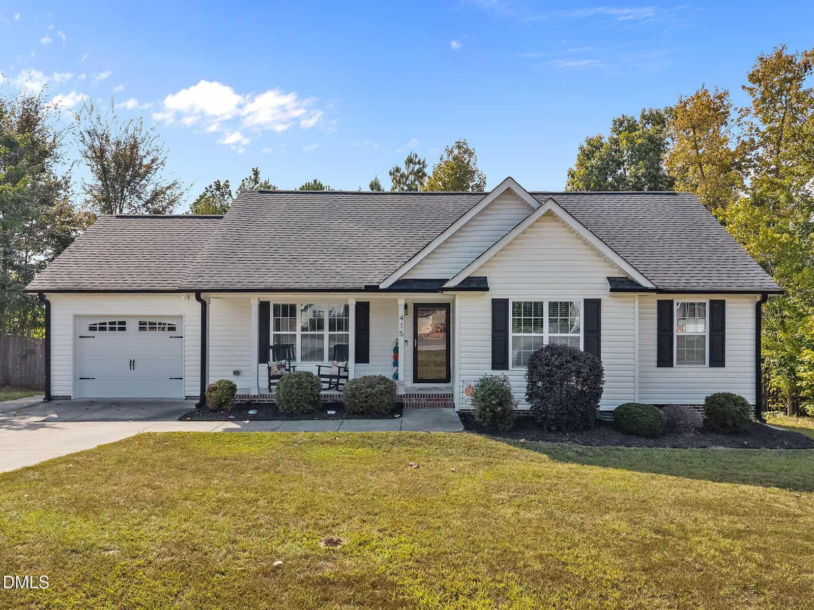 Charming single-story house in Stem at 415 Angus Place with white siding, black shutters, an attached garage on the left, a covered front porch with chairs, trimmed bushes, and a well-maintained front lawn under a partly cloudy sky.