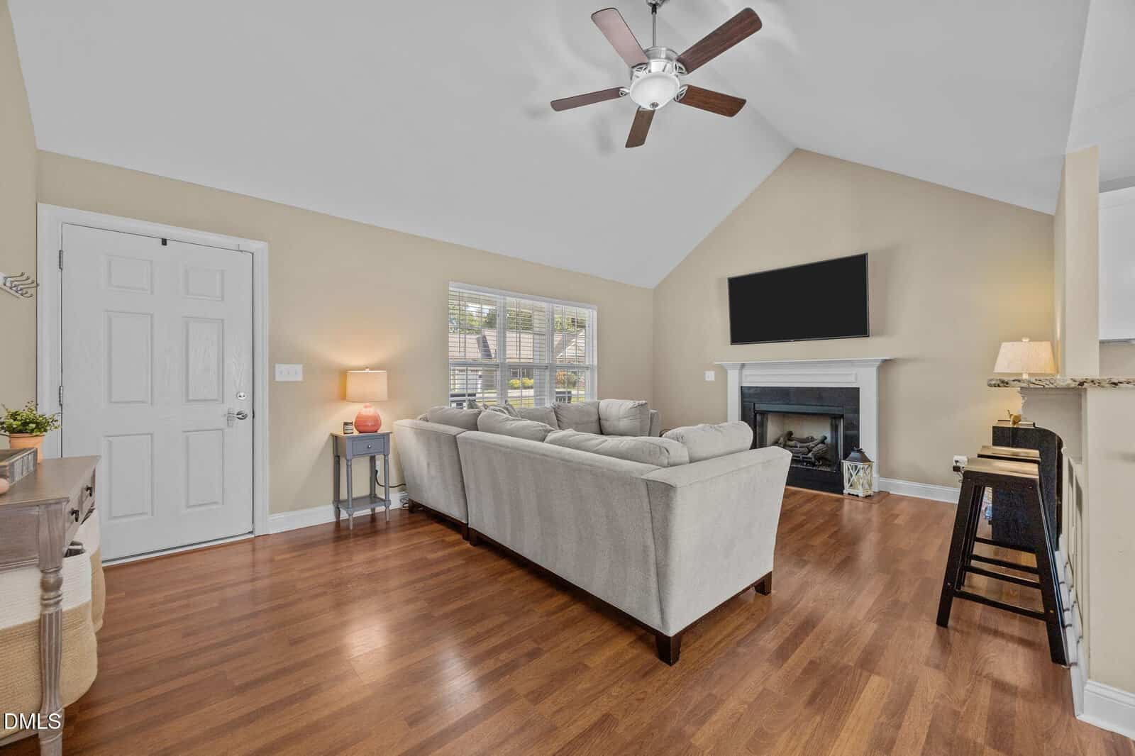 Living room with hardwood floors, beige walls, a gray sectional sofa, a large TV above a fireplace, ceiling fan, side tables with lamps, and windows letting in natural light. Door and entryway visible on the left.