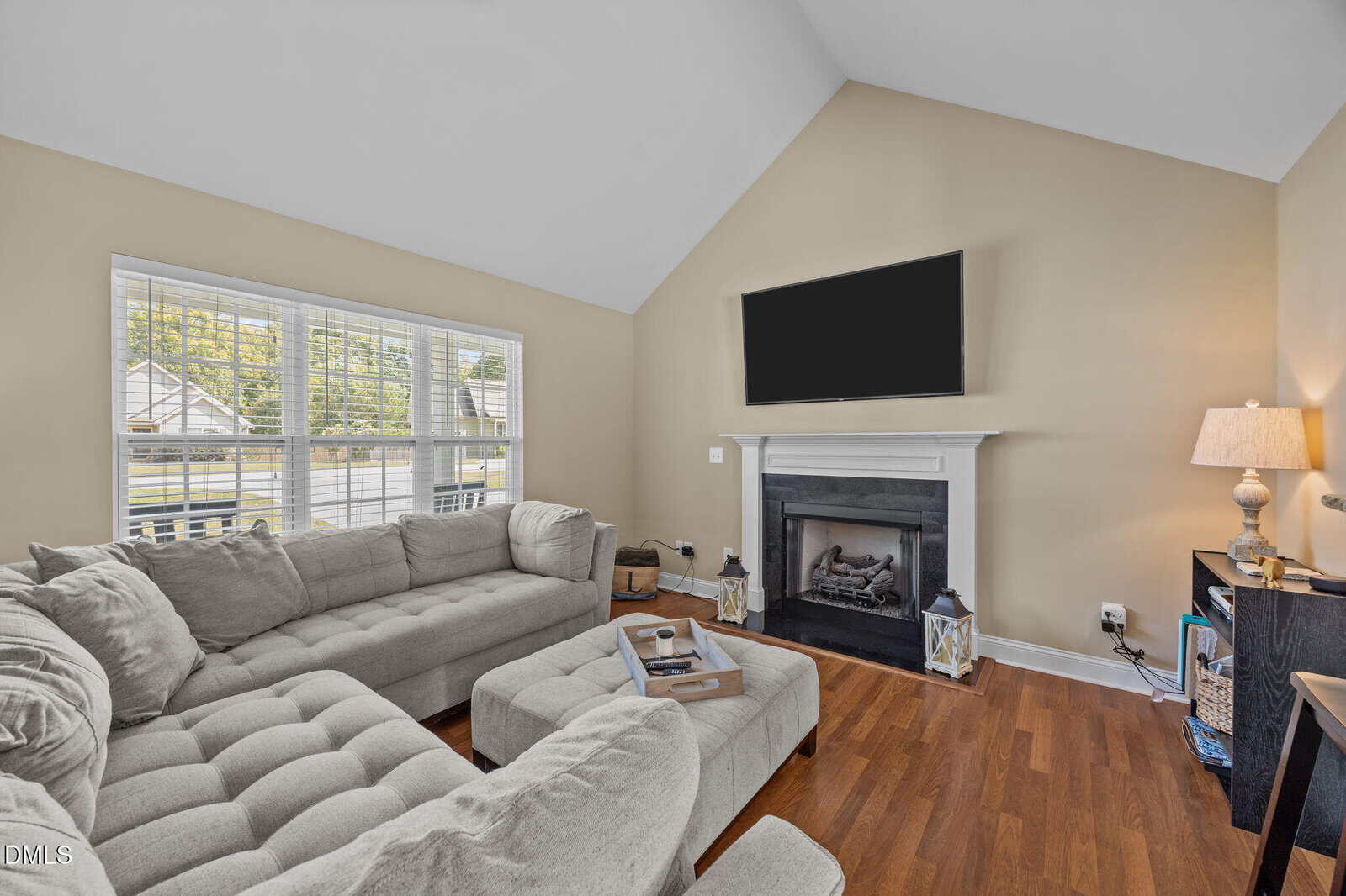 A living room with tan walls, wood floors, a sectional sofa, an ottoman, a fireplace with a TV above it, a table lamp, shelves, and a large window letting in natural light.