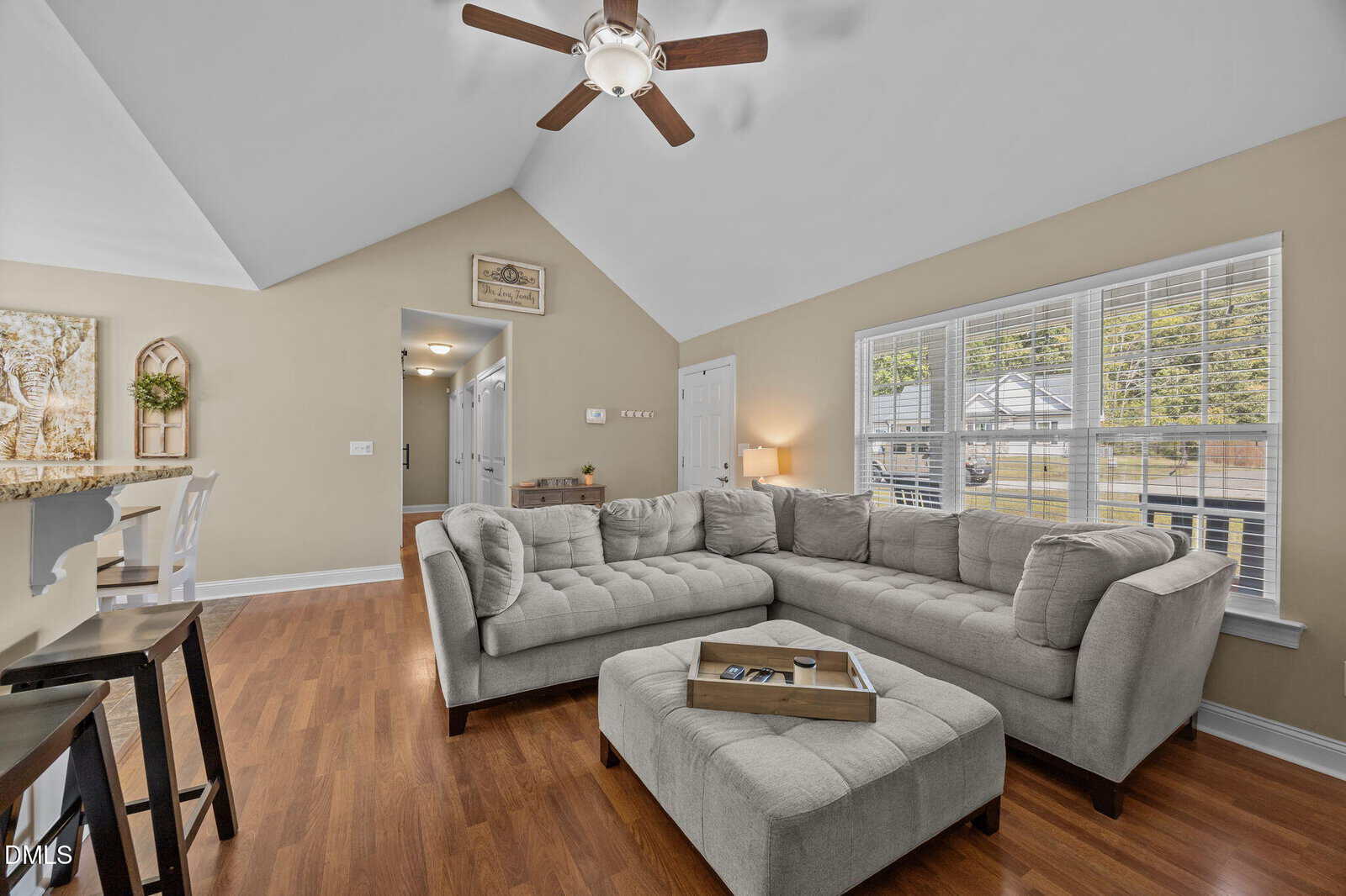 A living room with a gray L-shaped sectional sofa, matching ottoman, wooden floors, a ceiling fan, neutral walls, and large windows letting in natural light. A hallway and part of a kitchen are visible in the background.