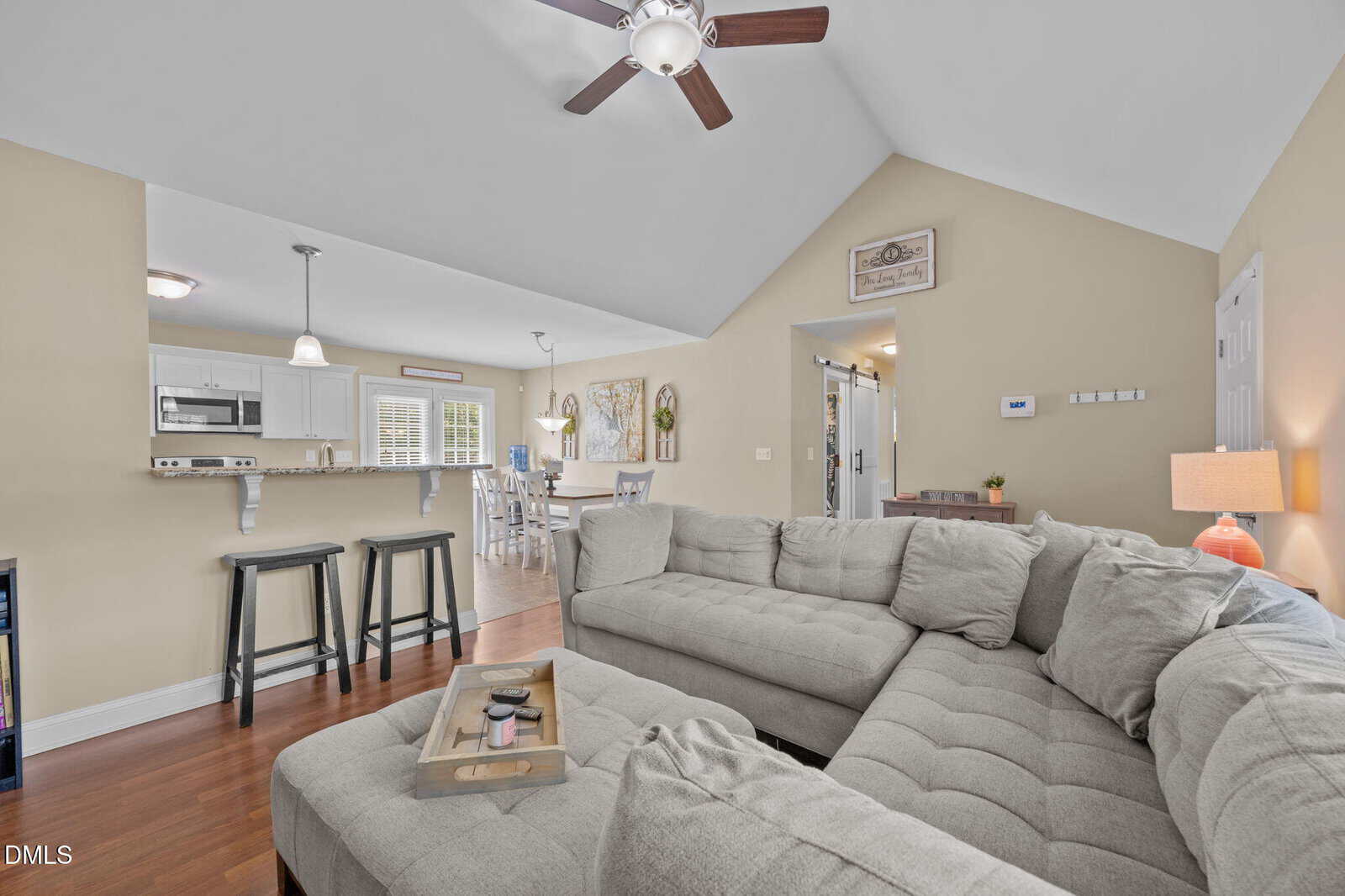 A spacious living room with a gray sectional sofa, ceiling fan, wooden floors, and beige walls. The open layout leads to a kitchen with white cabinets, a breakfast bar, and two black barstools.