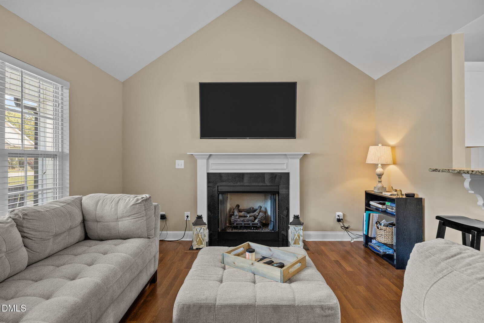 A living room with beige walls, a mounted flat-screen TV above a white fireplace, gray sectional sofas, a cushioned ottoman with a tray, wood flooring, a window with blinds, and a small lamp on a cabinet.