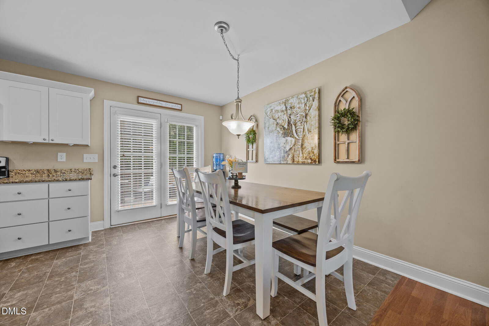 A kitchen dining area features a wooden table with four white chairs, a hanging light above, beige walls with framed art and a window decor piece, and a door with blinds leading outside. White cabinets and granite countertops are visible.