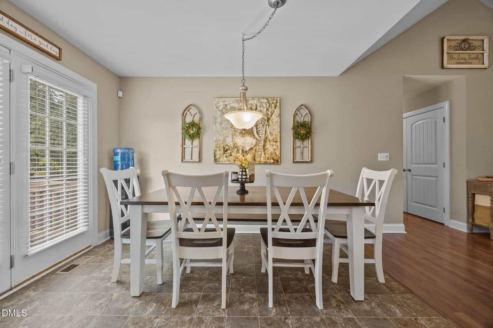 A dining area with a rectangular wooden table, six white chairs, a hanging light fixture, wall art, and decorative greenery. The room has beige walls, tile flooring, and a water dispenser by glass doors.