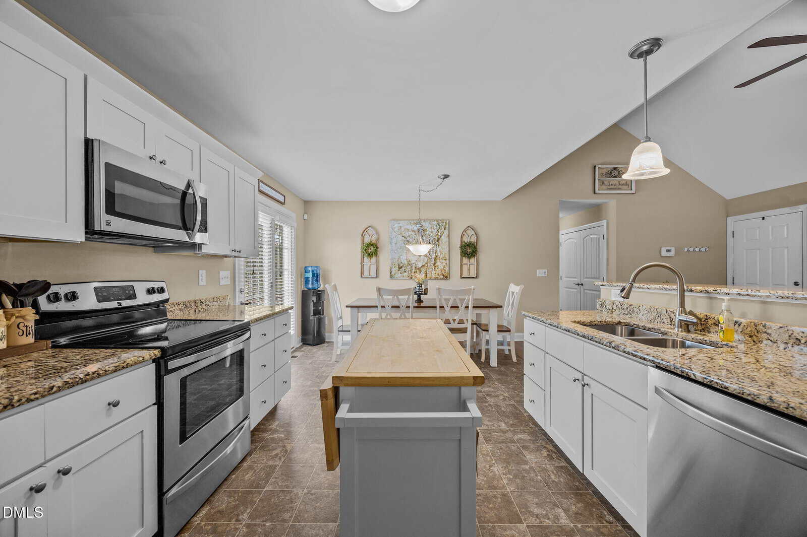 Modern kitchen with white cabinets, granite countertops, stainless steel appliances, a wooden island, and a dining area in the background with a round table, chairs, wall mirrors, and decorative plants.