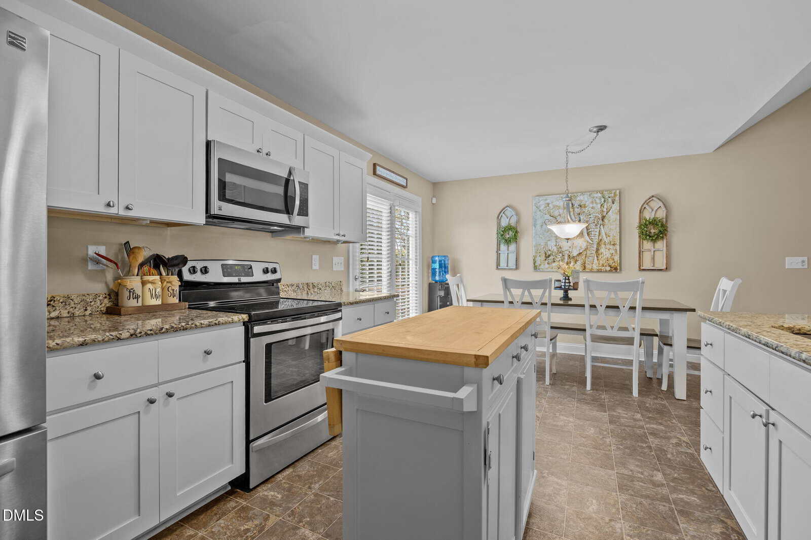 Modern kitchen with white cabinets, stainless steel appliances, a small center island with a wooden top, and a dining area with a table and chairs next to a large window with blinds.