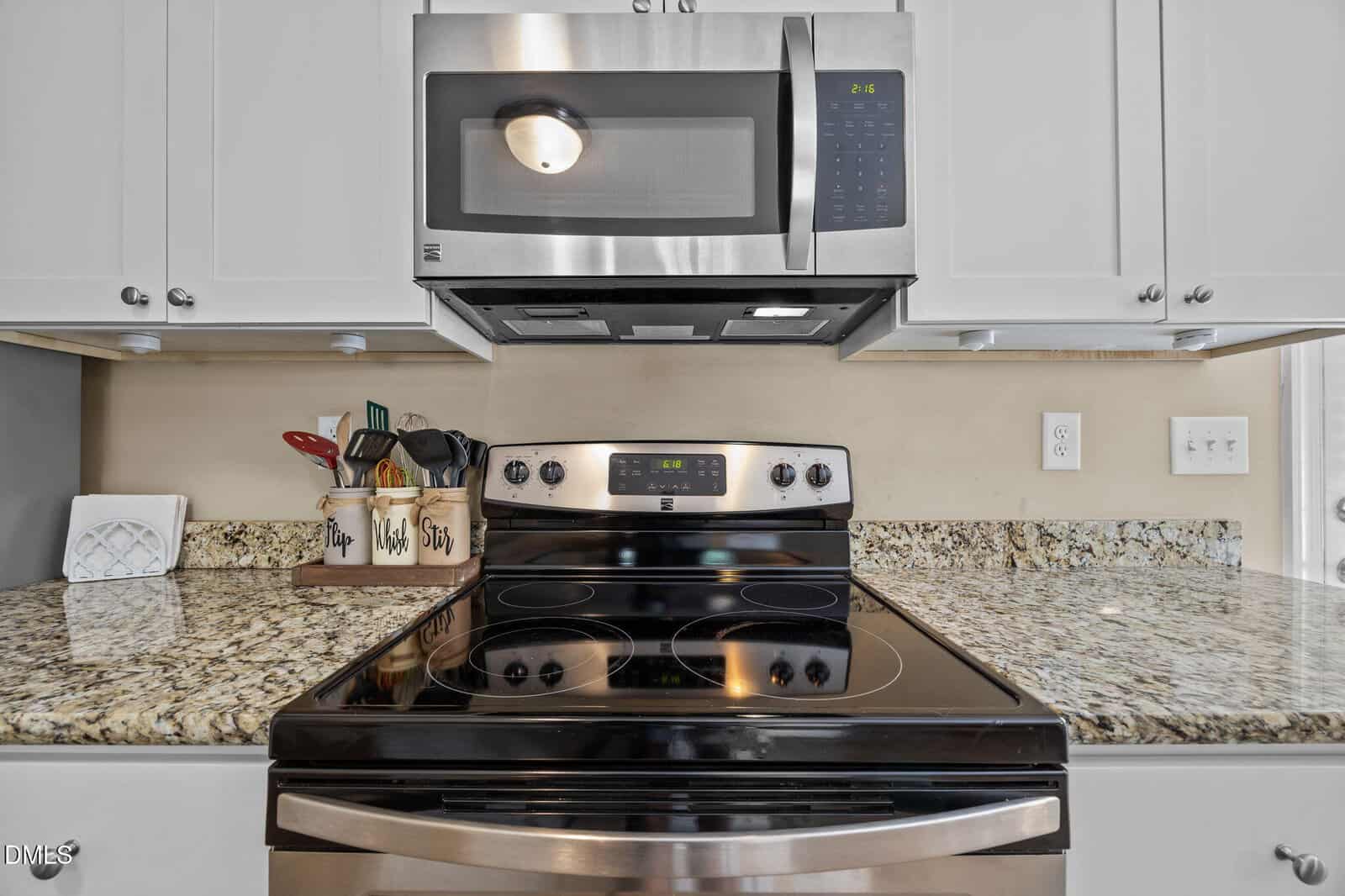 Stainless steel stove and microwave on granite countertop, surrounded by white cabinets. Utensils, spices, and dishes are on the left. The wall outlet and light switch are on the right.